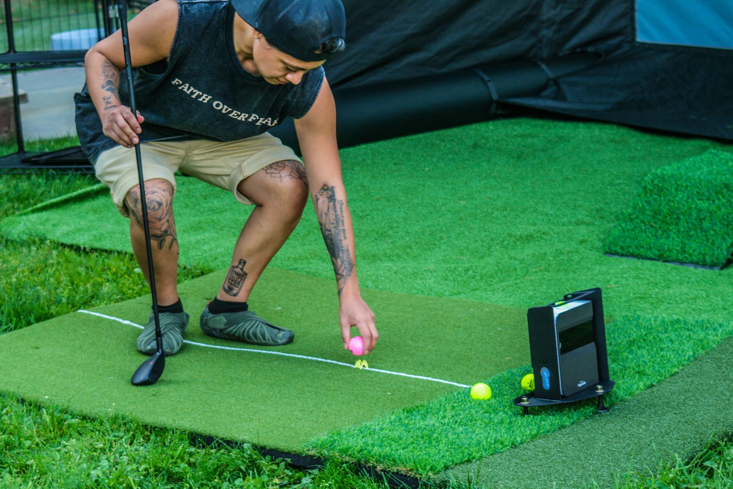 A person with tattoos, wearing a black sleeveless shirt, beige shorts, black shoes, and a black cap, is crouched down on green artificial turf, preparing to putt a pink golf ball. There are three other balls on the turf and a smartphone on a black stand nearby.