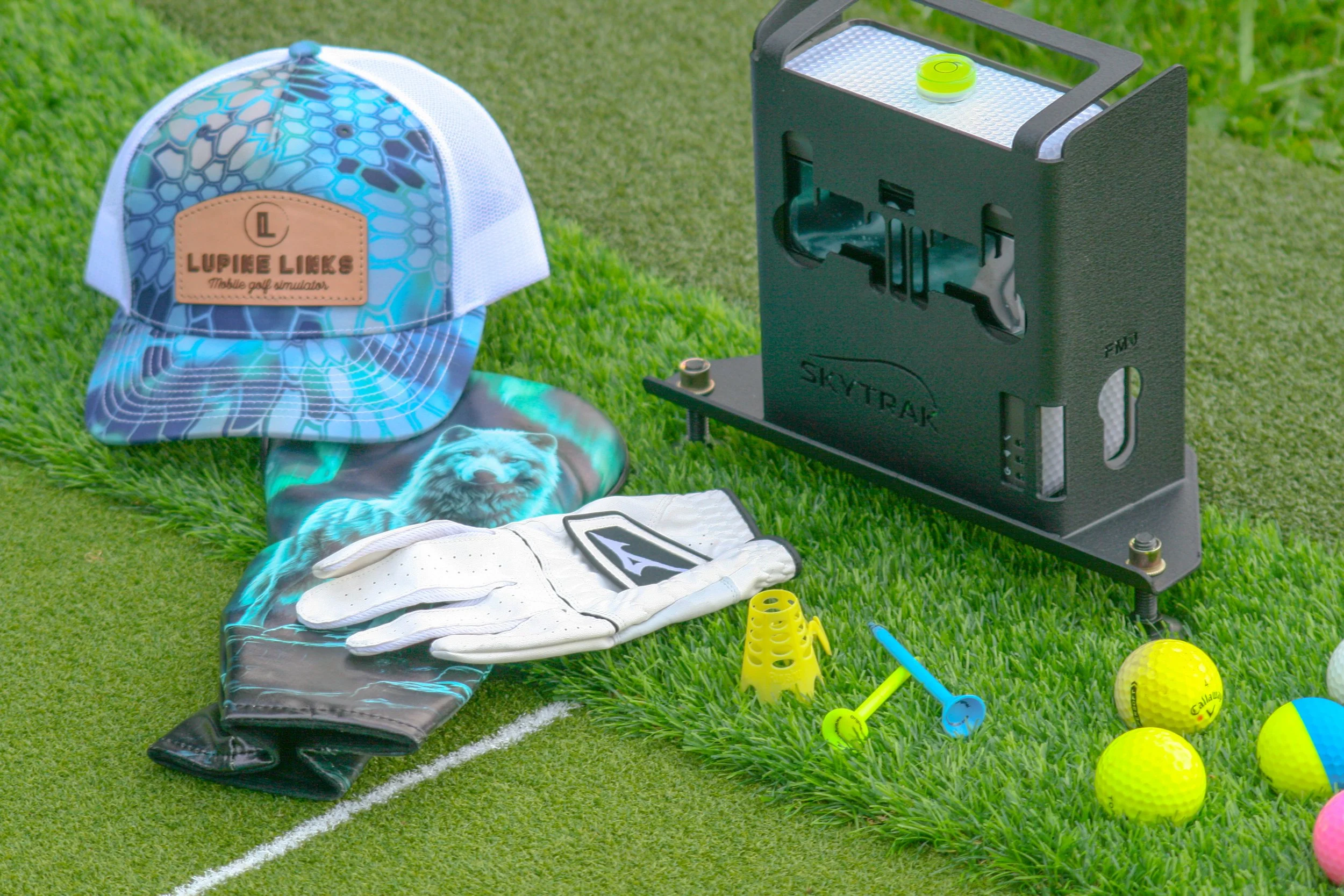 Golf equipment and accessories arranged on a grassy outdoor surface, including a colorful baseball cap, golf glove, golf balls, a tee, a Slice golf ball marker, and a golf simulator machine.