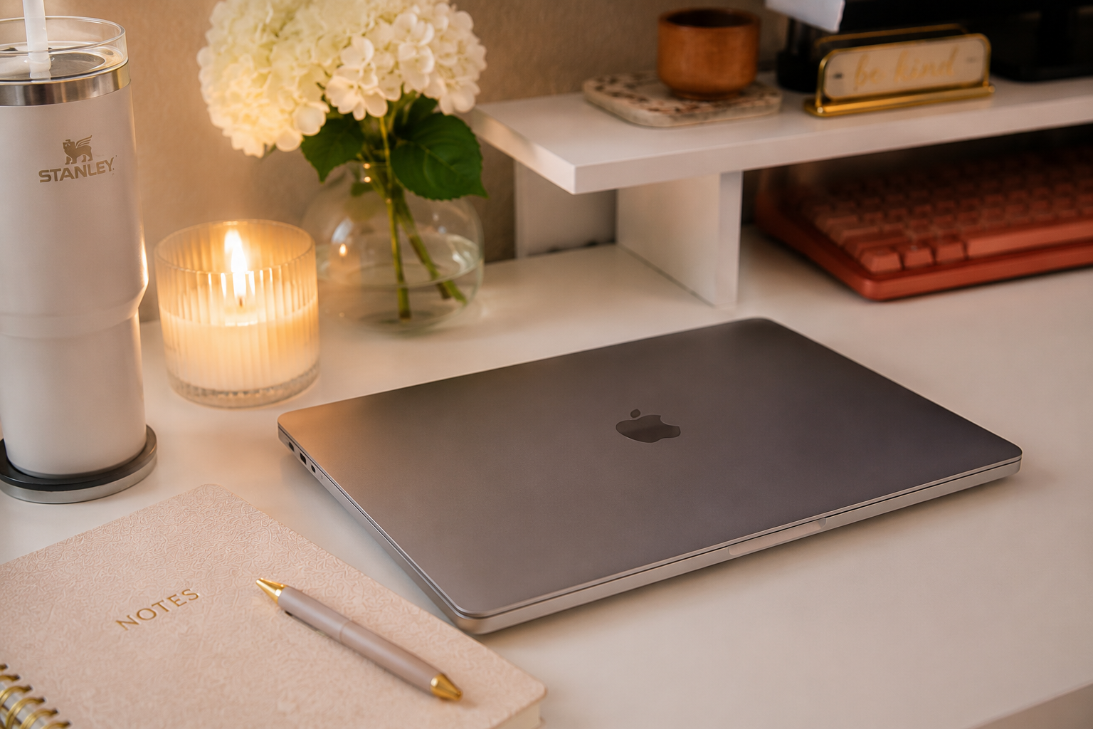 A tidy workspace with a closed MacBook, a notebook titled 'NOTES' with a gold pen on top, a lit candle, a small glass vase with white flowers, a candle holder, a white Stanley brand water bottle, and a red keyboard on a white desk with a shelf above holding a wooden container and a sign that says 'be kind'.