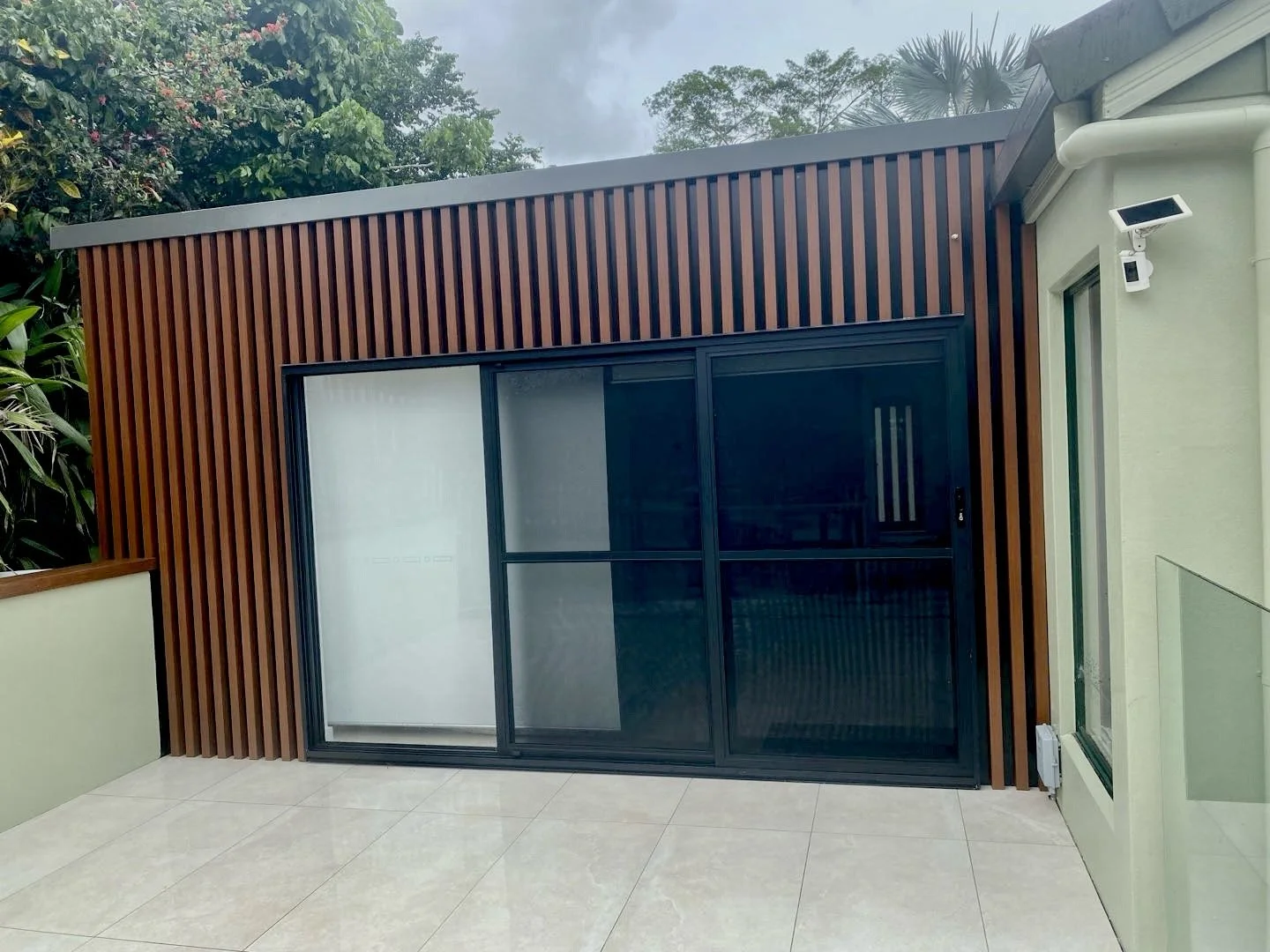 Exterior view of a modern building with a sliding glass door and wooden slat wall, surrounded by greenery and a tiled patio.
