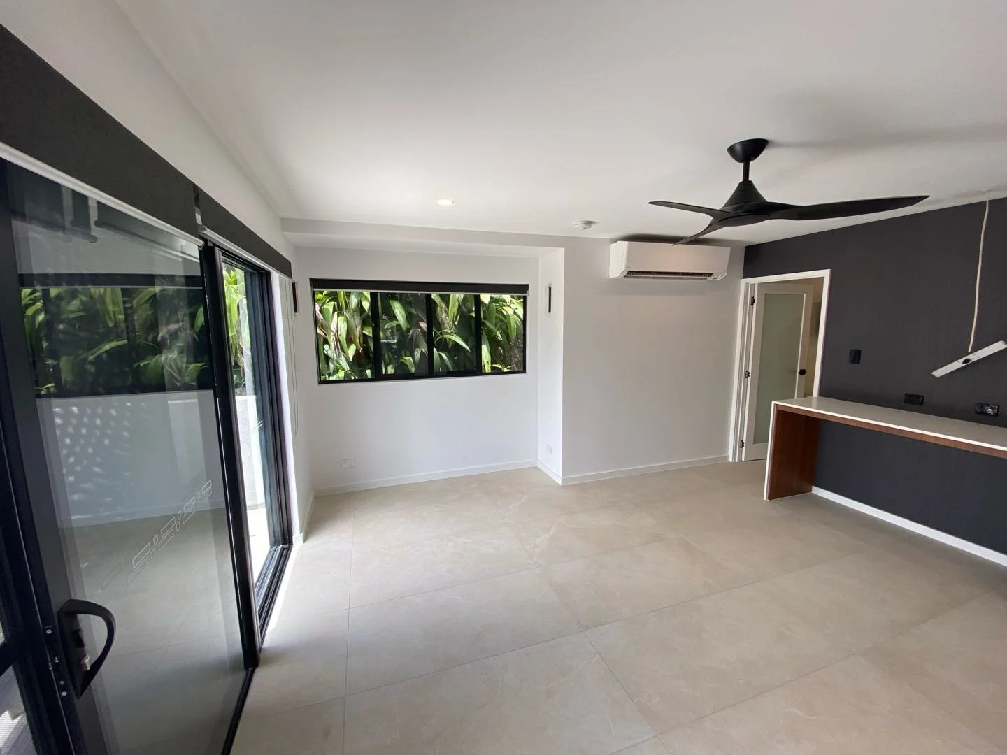Empty modern living room with sliding glass doors, black window frames, a ceiling fan, air conditioner, and a dark accent wall with a white desk surface.