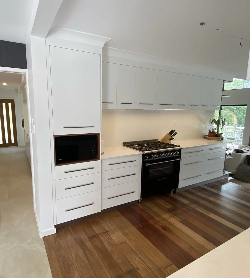 Modern white kitchen with cabinets, a black oven and stovetop, and wood flooring, with a view of a living area and outdoor space.