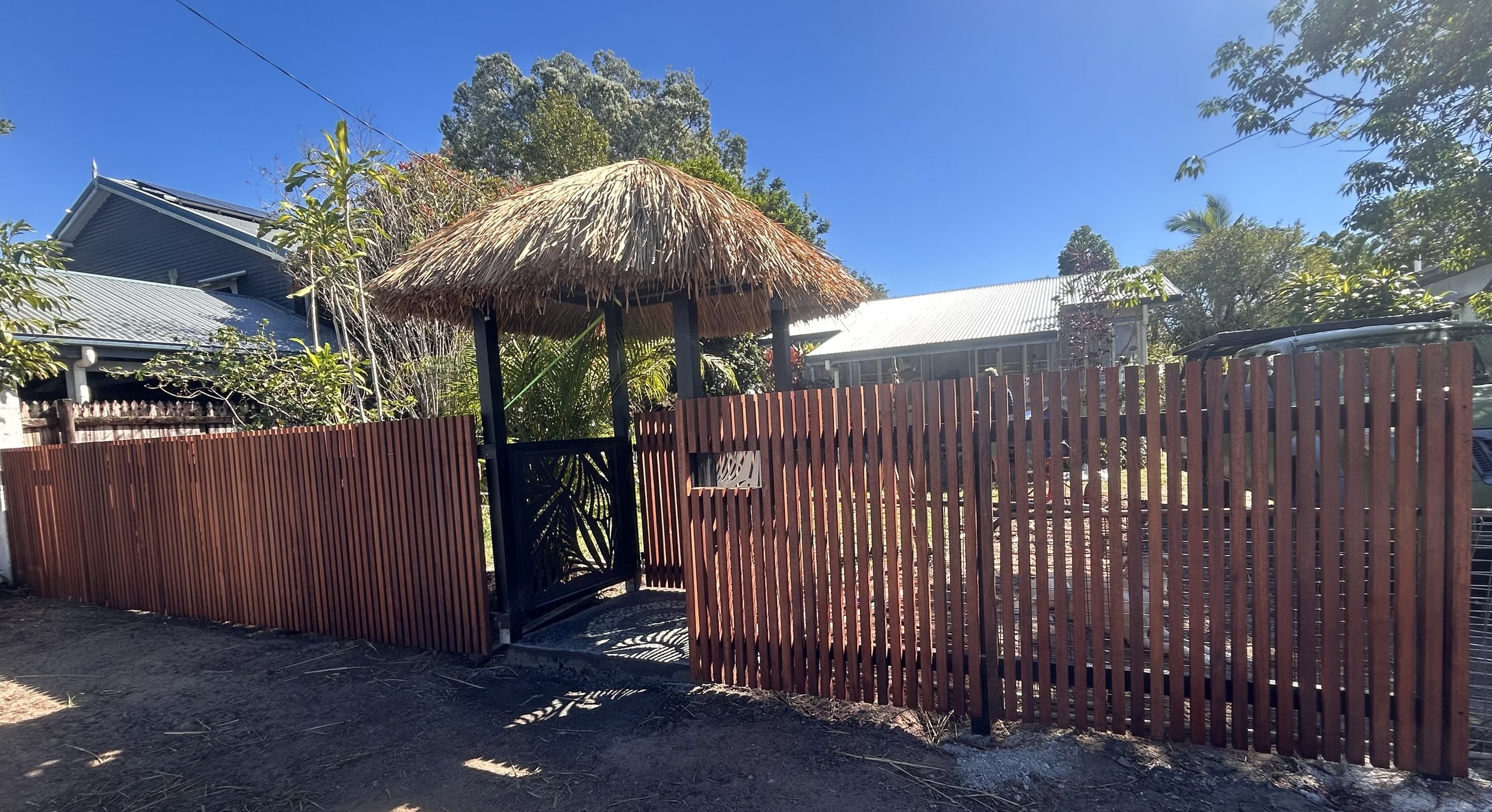 A backyard with a wooden fence, gate, and a thatched-roof hut surrounded by trees and neighboring houses under a clear blue sky.