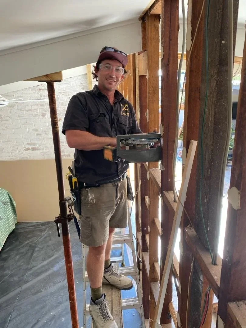 A man wearing safety glasses, a baseball cap, and work clothes, standing on a ladder and working on a house wall framework during renovations.