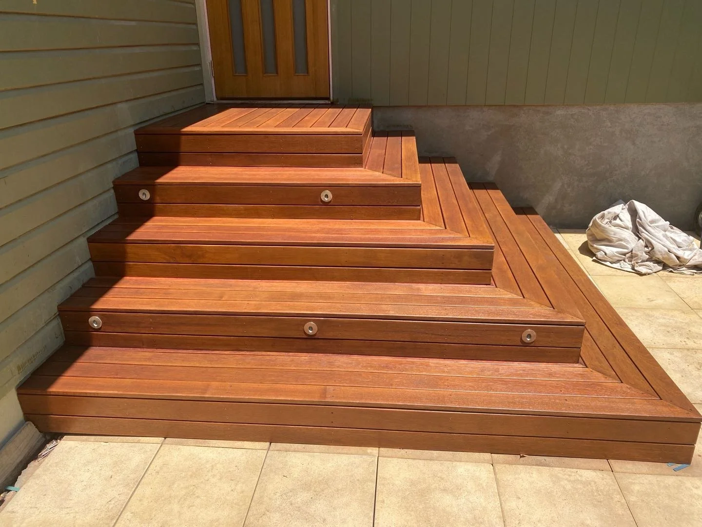 Wooden deck stairs leading to door, with a small landing, installed on a tiled patio with some crumpled cloth on the side.