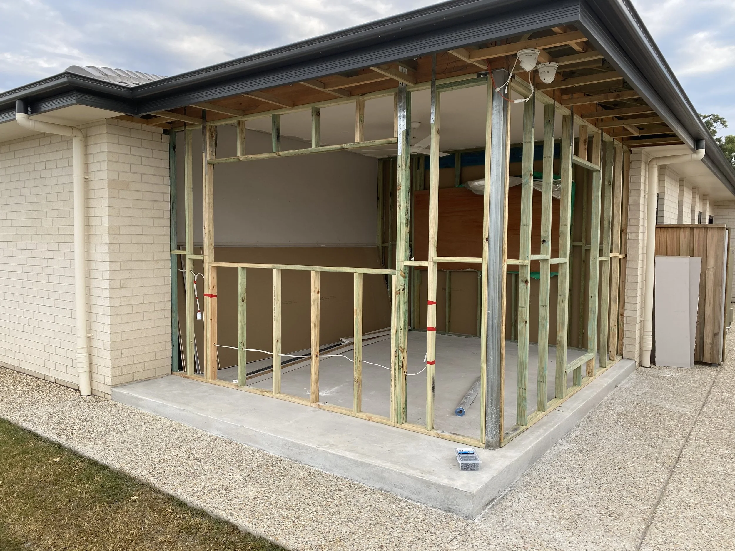 Front view of a house under renovation with exposed wooden framing for an extension or porch, with side brick walls, a concrete foundation, and a garaged roof under cloudy sky.
