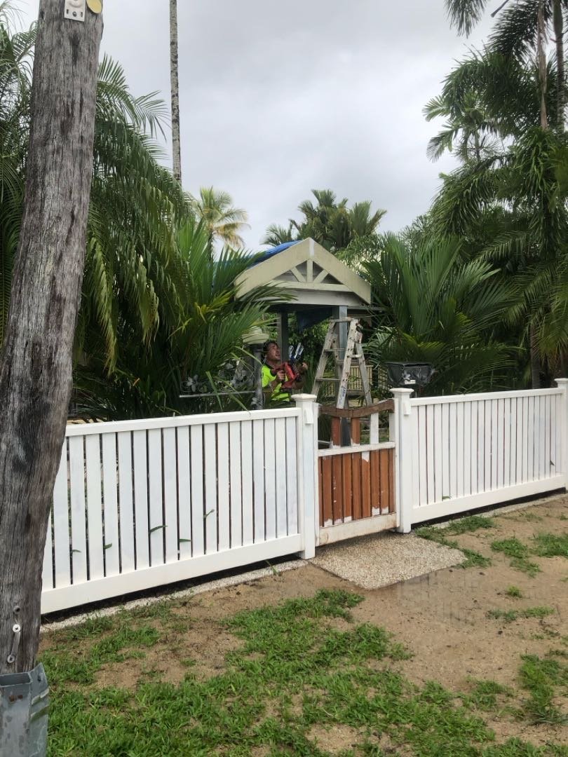Person working on a house roof with tools, ladder, and worker gear, behind a white fence surrounded by palm trees.