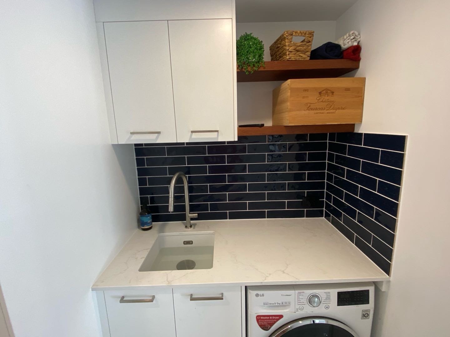 Small kitchen area with white cabinets, black subway tile backsplash, a white marble-look countertop, a small undermount sink, a soap dispenser, open wooden shelves with decorative items including a green plant, a wooden wine box, and folded clothes, and an LG washing machine underneath the counter.