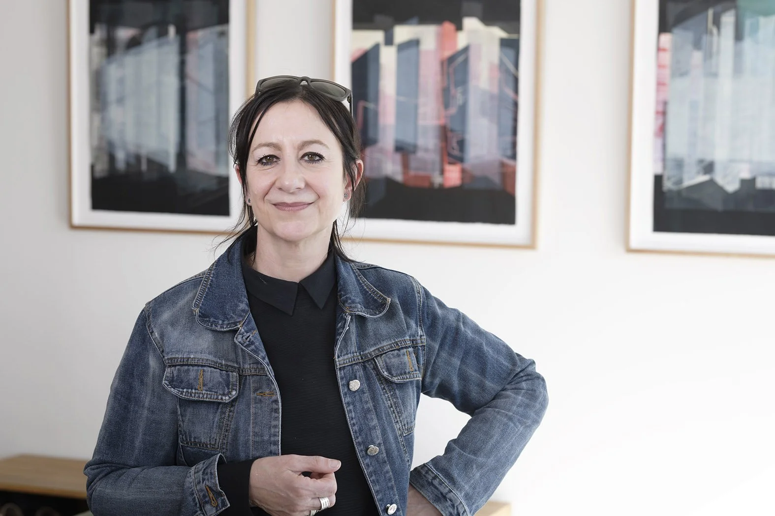 A woman smiling and standing indoors in front of framed artwork, wearing a dark turtleneck, denim jacket, and sunglasses on her head.