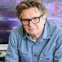 Artist John Waller with glasses and gray hair wearing a denim shirt, sitting in front of an abstract landscape with a colorful background.