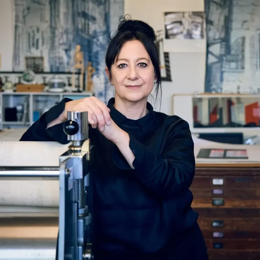 Artist and printmaker Cat Poljski  with dark hair in a messy bun, wearing a black top, stands in a creative workspace or studio, leaning on a large print press , with an abstract mural, shelves, and art supplies in the background.