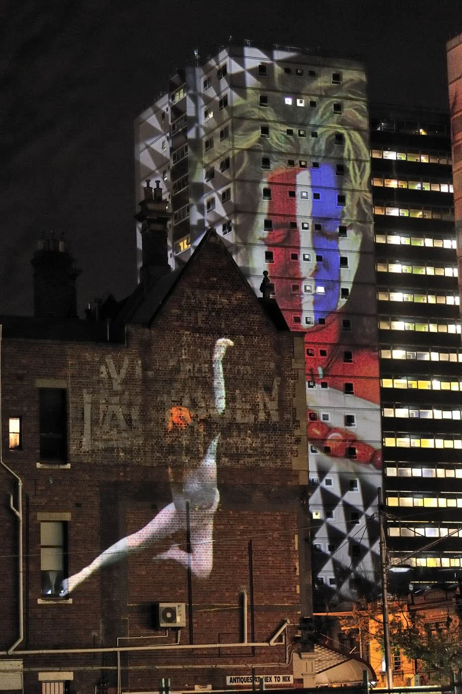 Nighttime city scene with a large projection of a classical painting on a brick wall and a high-rise building in the background. The projection features a woman with a blue and red garment, and there are bright lights illuminating the area.