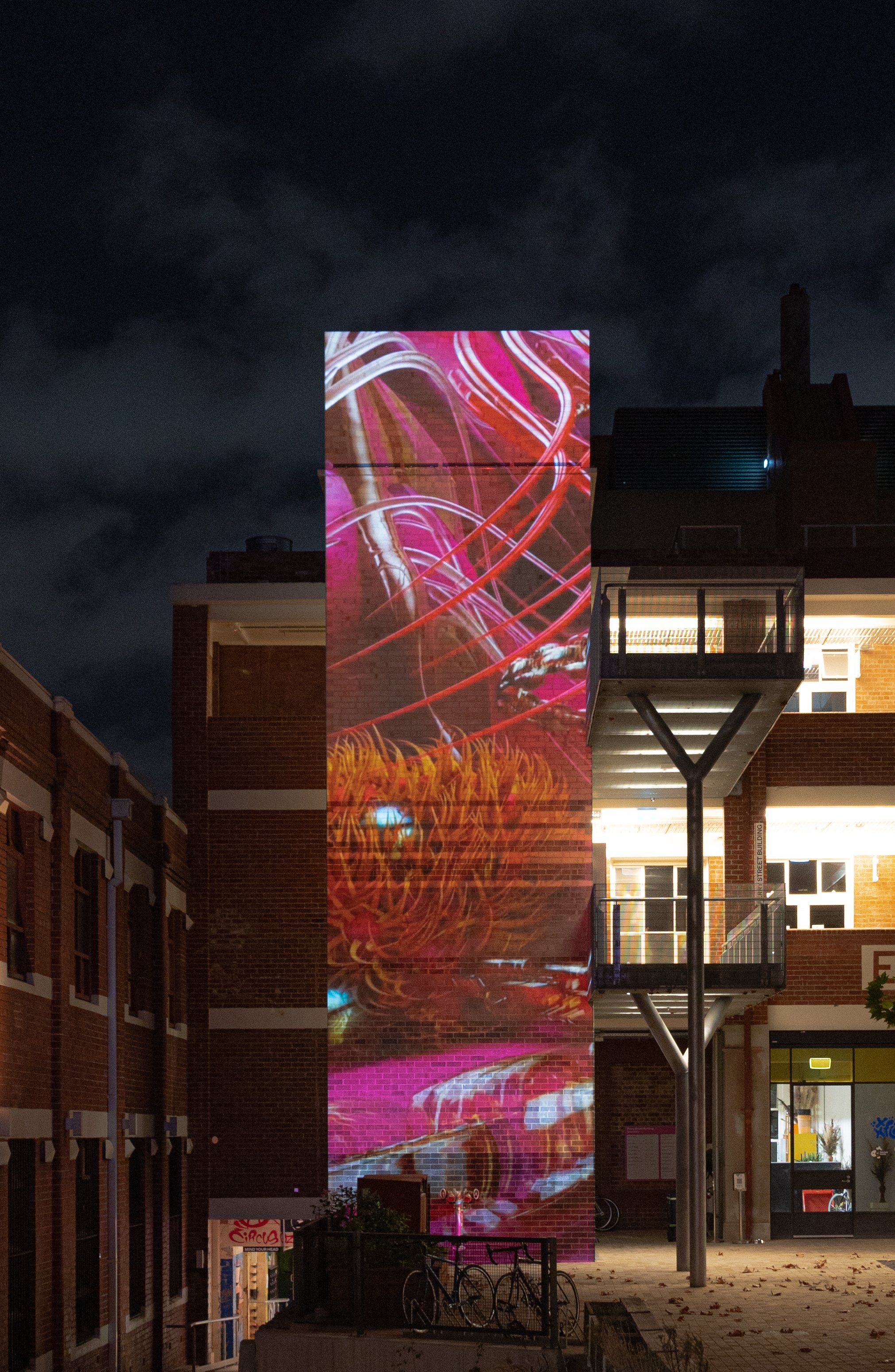Night scene showing a brick building with a large vertical digital projection of colorful abstract art on it. The projection displays swirled patterns in red, pink, white, and orange. There is a metal fire escape structure with stairs and a platform in front of the building, and bicycles parked at the bottom. The area is illuminated with outdoor lights, and there are some fallen leaves on the ground.