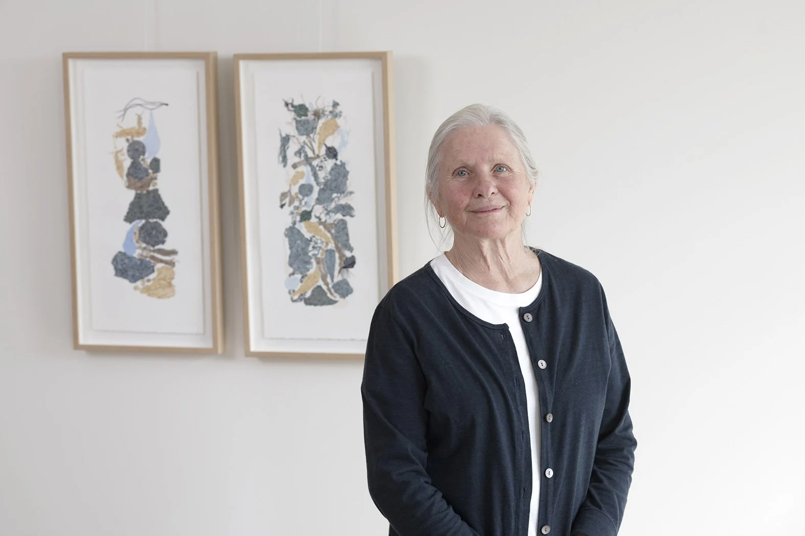An artist with gray hair, wearing a black cardigan and white top, standing in front of two framed abstract art pieces on a white wall.