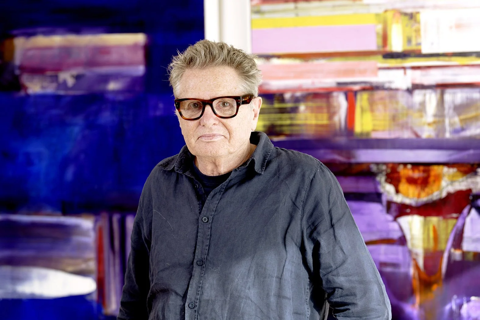 An artist with gray hair, wearing glasses and a dark shirt, standing in front of colorful abstract artwork.
