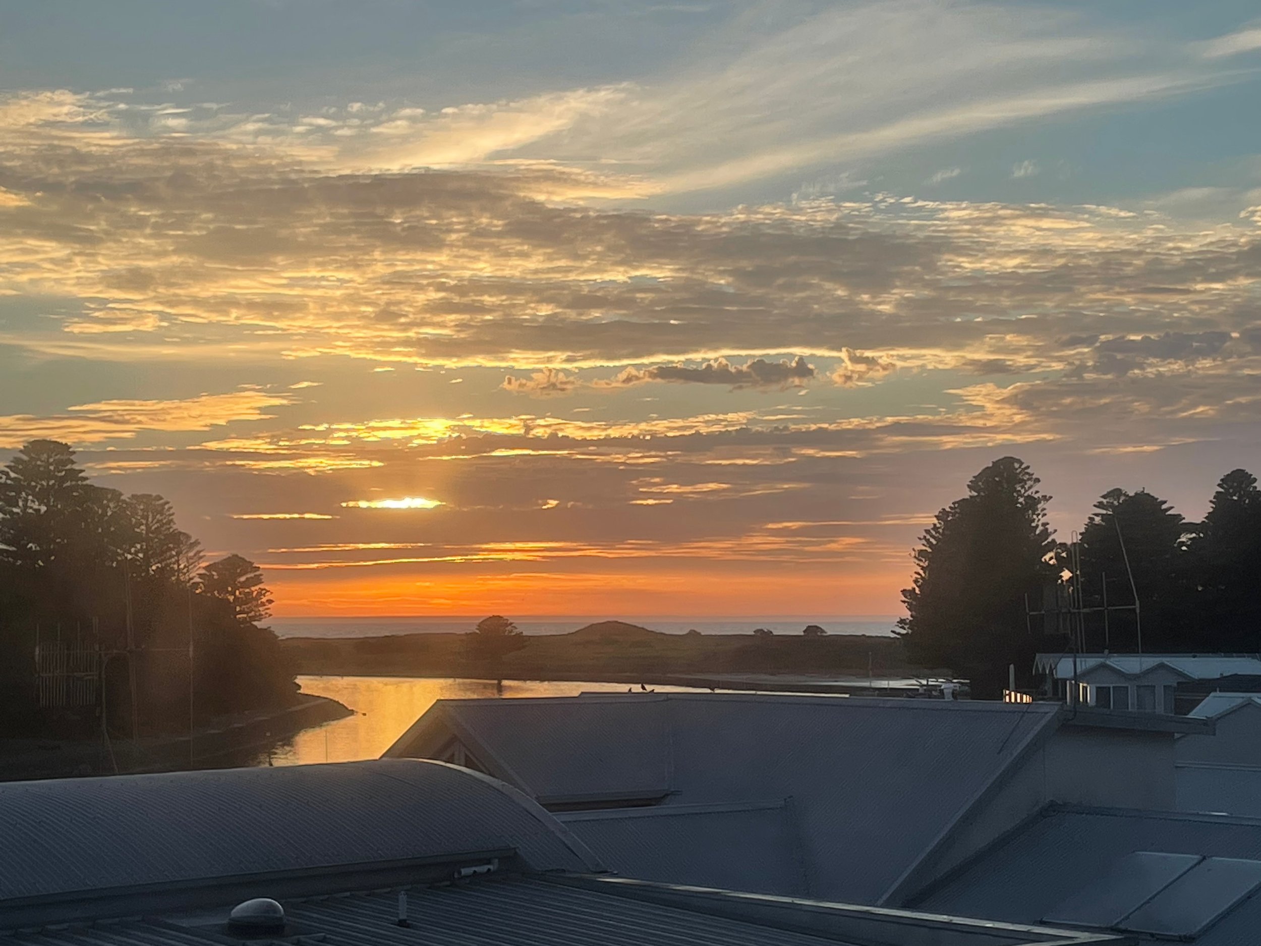 A sunset view over a body of water with trees silhouetted in the foreground, clouds scattered across the sky.