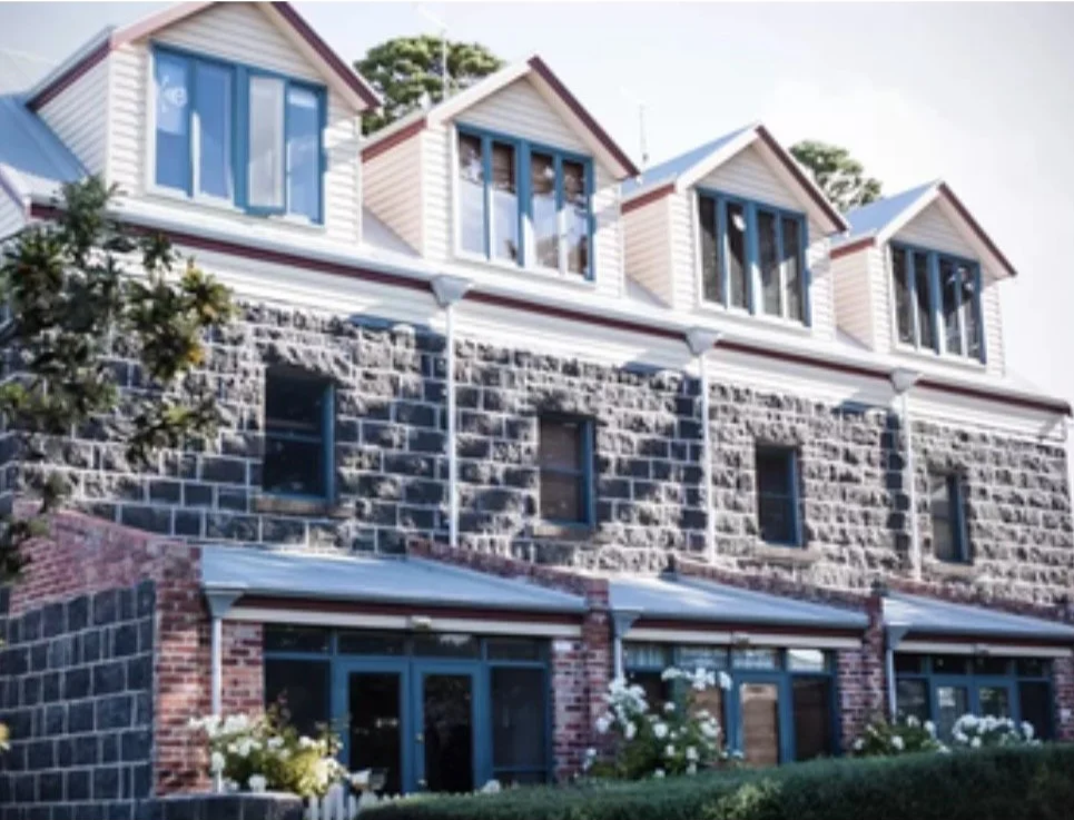 Three-story building with a brick and stone facade, featuring multiple windows and dormer windows on the top floor. A tree is visible on the left side.