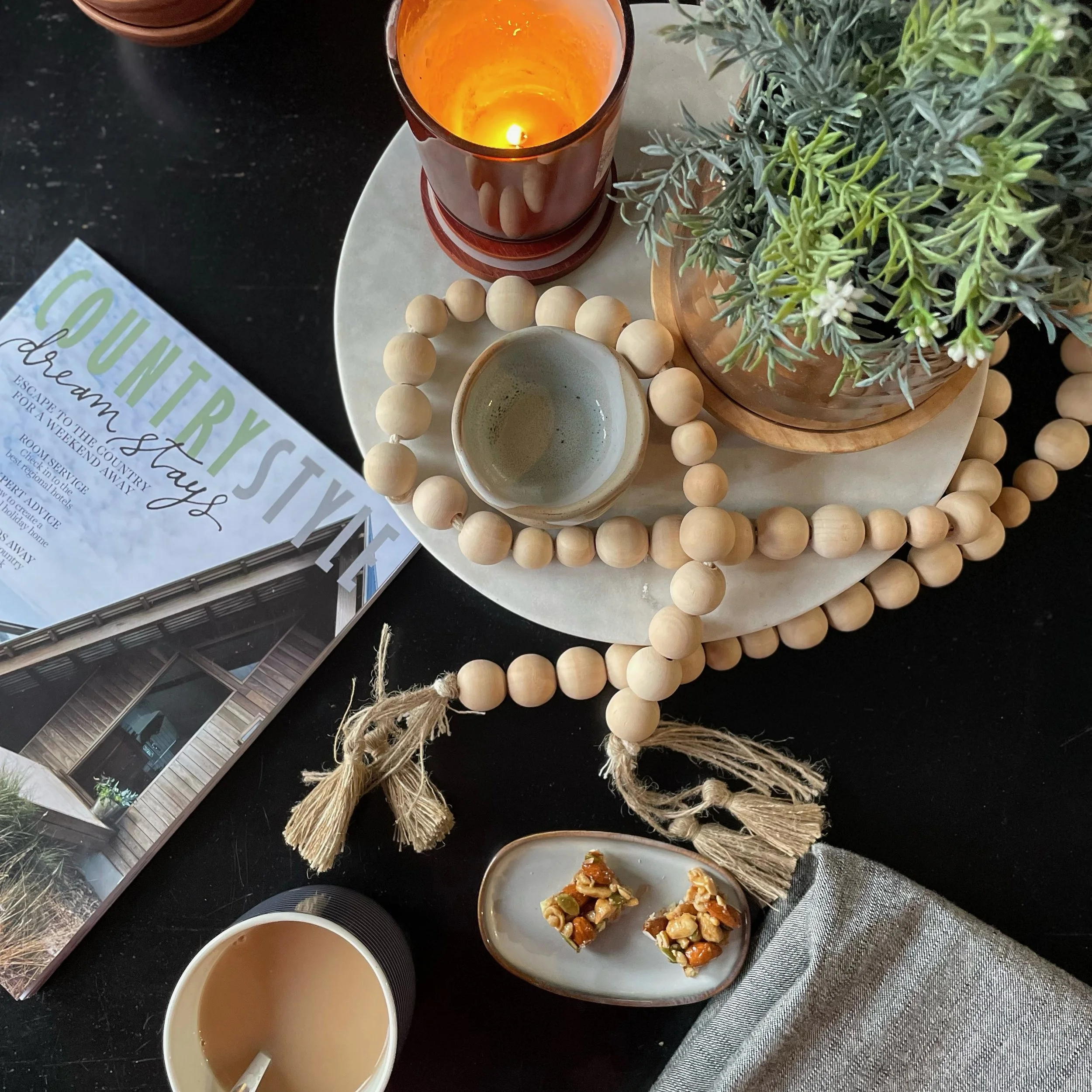 A table setup with a lit candle in a glass holder, a potted plant, a string of large wooden beads, a small bowl of empty water, two cups of coffee, a plate with a pastry topped with nuts and dried fruit, and a magazine with the headline 'Touristy'.