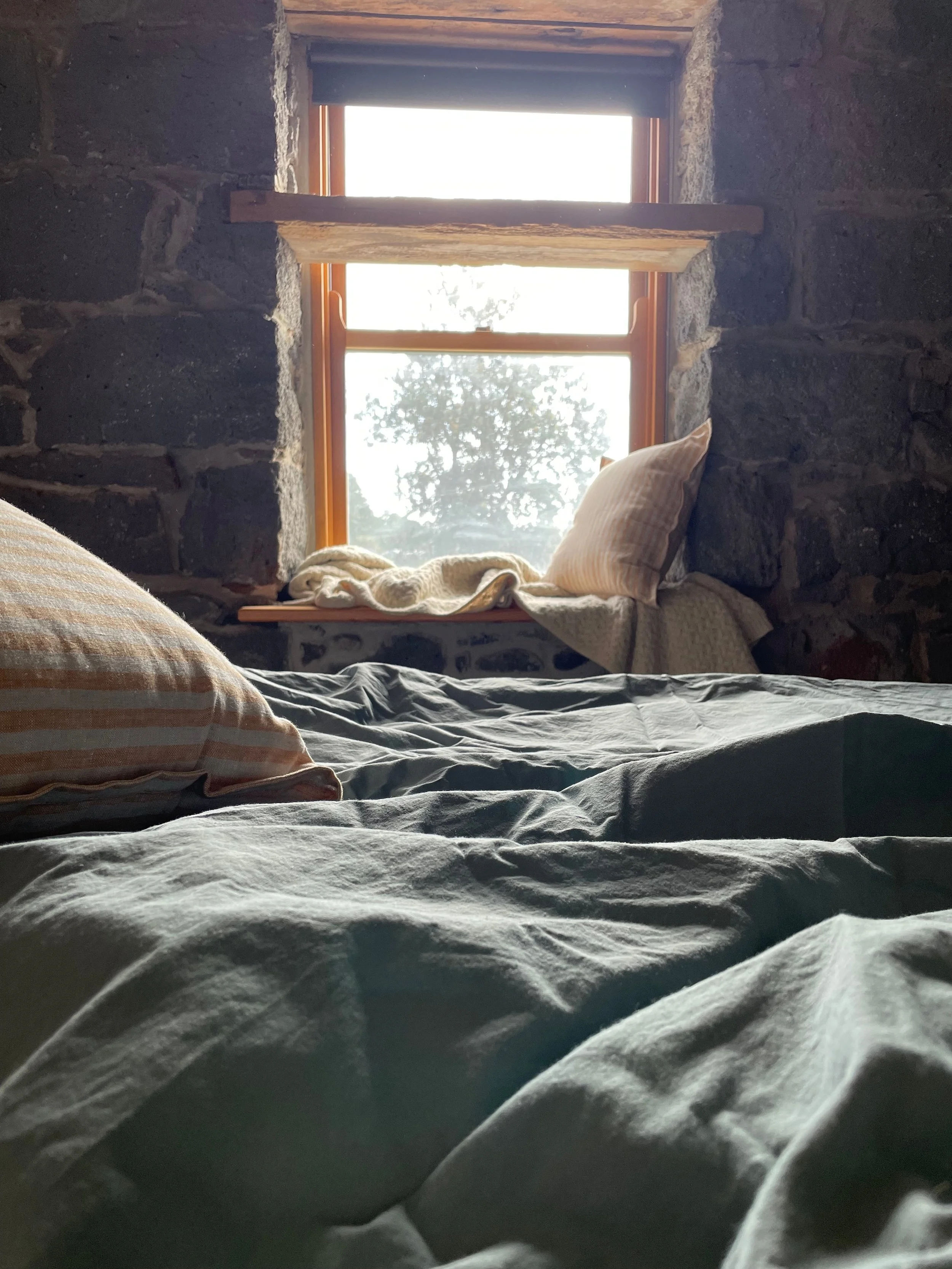 View from a bed looking towards a window with stone walls on either side and a pillow resting against the wall. The window provides natural lighting, revealing an outdoor tree.