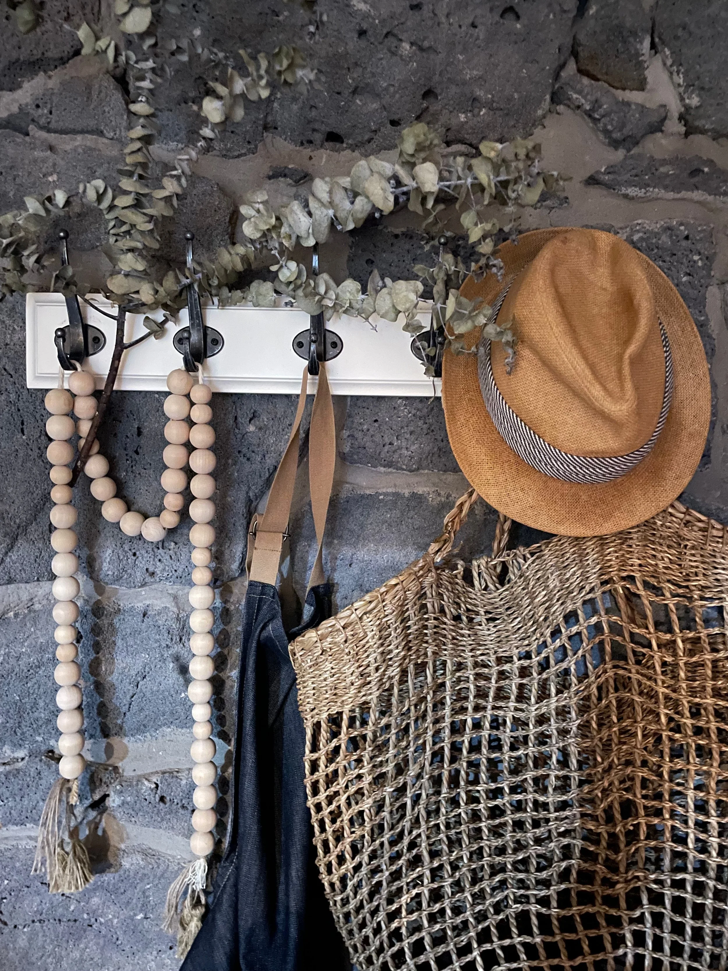 Close-up of a stone wall with hanging items, including a straw hat with a black and white striped band, a woven basket, a rope, a decorative beaded necklace, and dried branches or leaves.