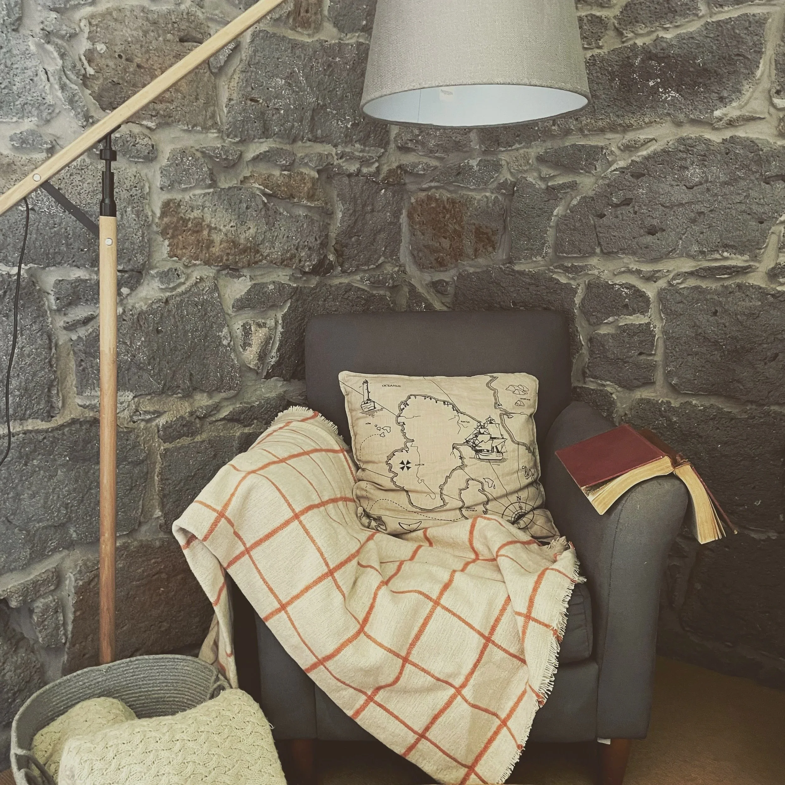 A corner of a room with a rustic stone wall. A beige lampshade hangs from the ceiling. There is a black armchair with a beige blanket featuring orange stripes. On the floor, there are wicker baskets and a large cloth with a map and illustrations of s