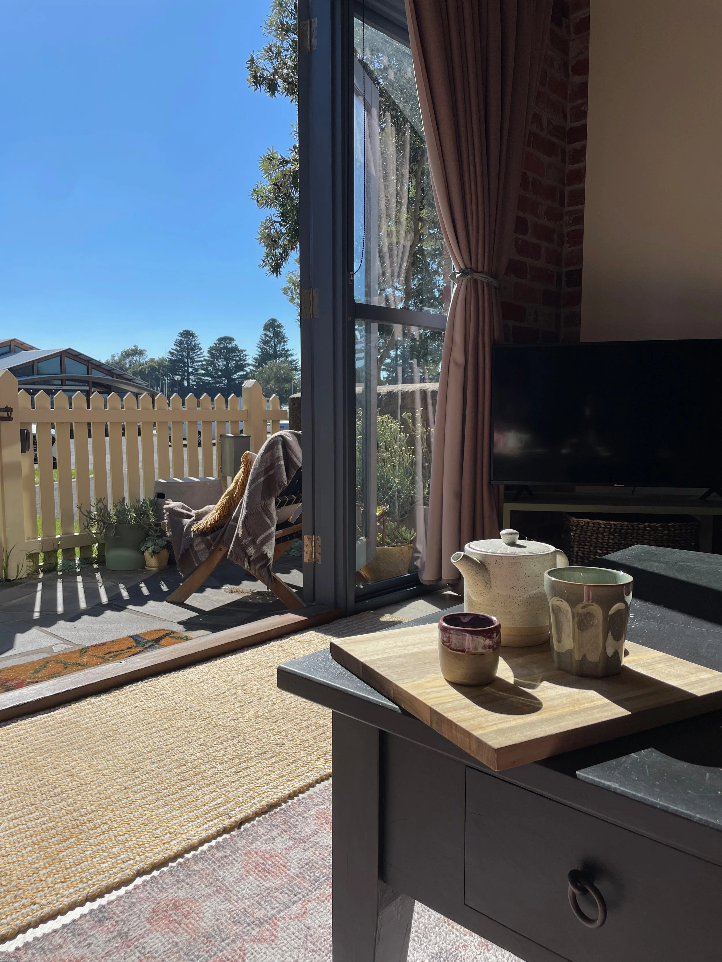 A living room with a sliding glass door opening to a sunny outdoor patio with a white picket fence and green trees. Inside, a table holds ceramic cups and a teapot, while a TV is positioned near a tan curtain tied back to reveal the garden outside.