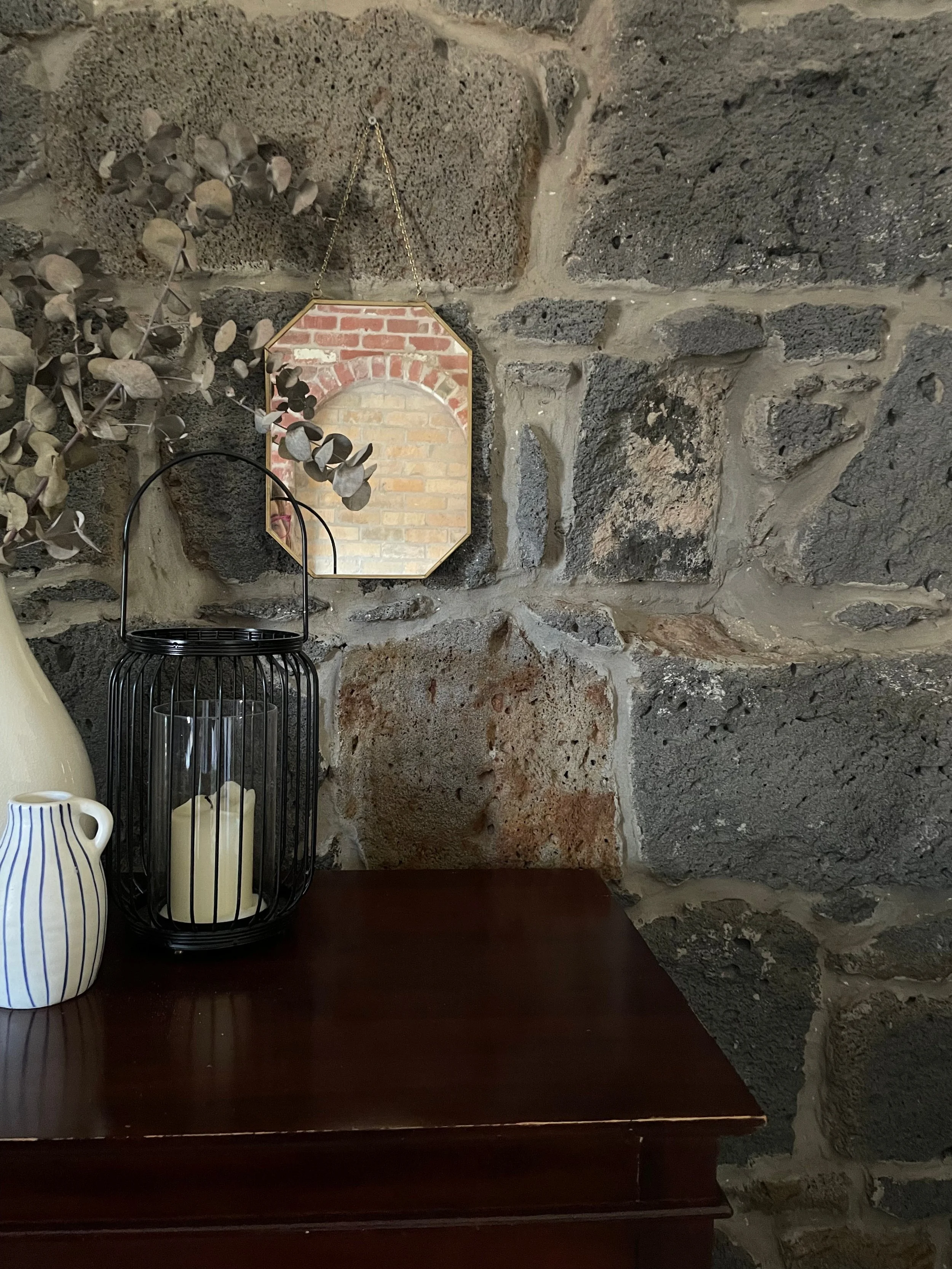 Decorative urn with candles, a small mirror hanging on a stone wall, and two striped vases on a wooden surface.