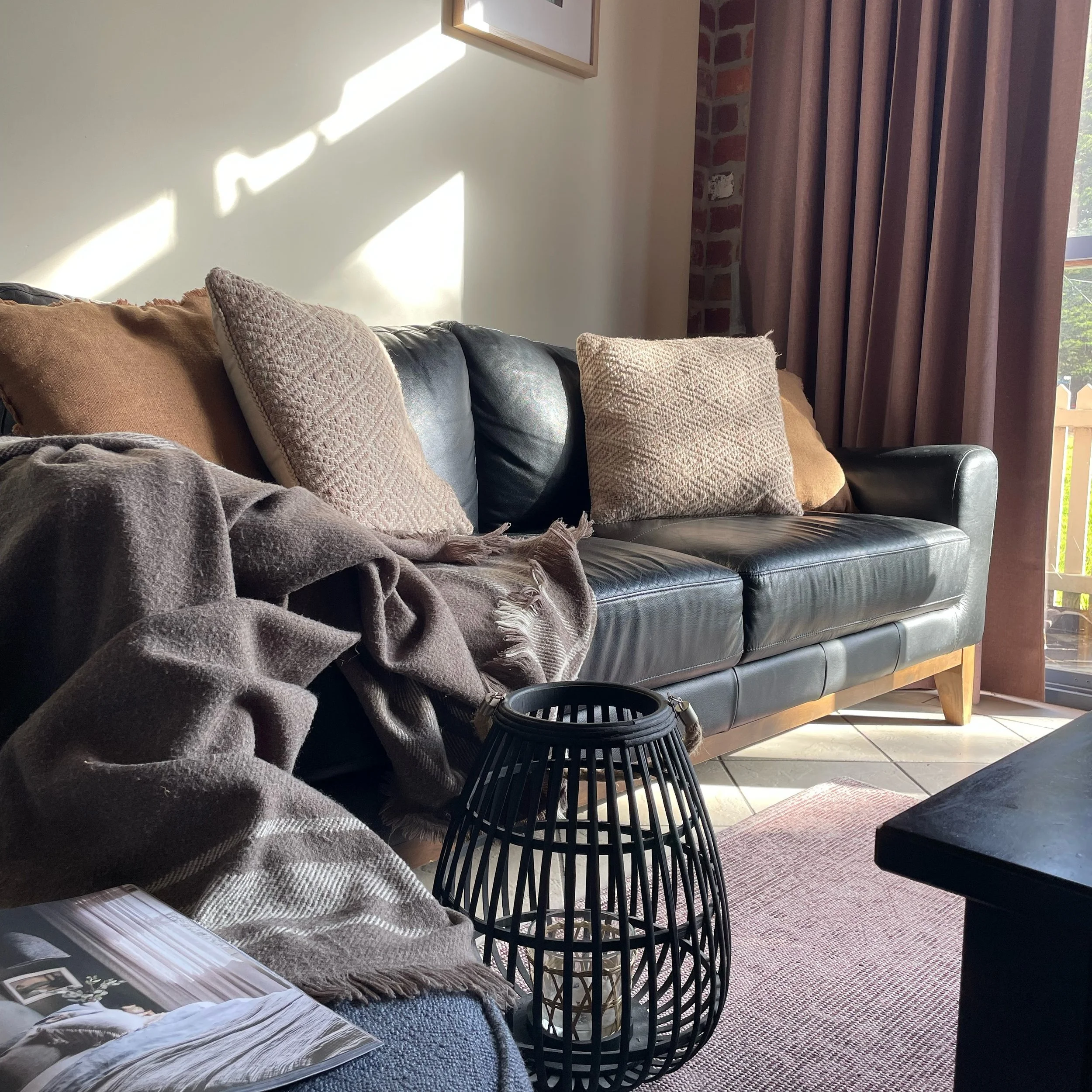 A cozy living room corner with a black leather sofa, beige and textured throw pillows, a blanket, and a magazine or book on the armrest. Sunlight streams through a window with beige curtains, illuminating the wall and part of an exposed brick wall. T