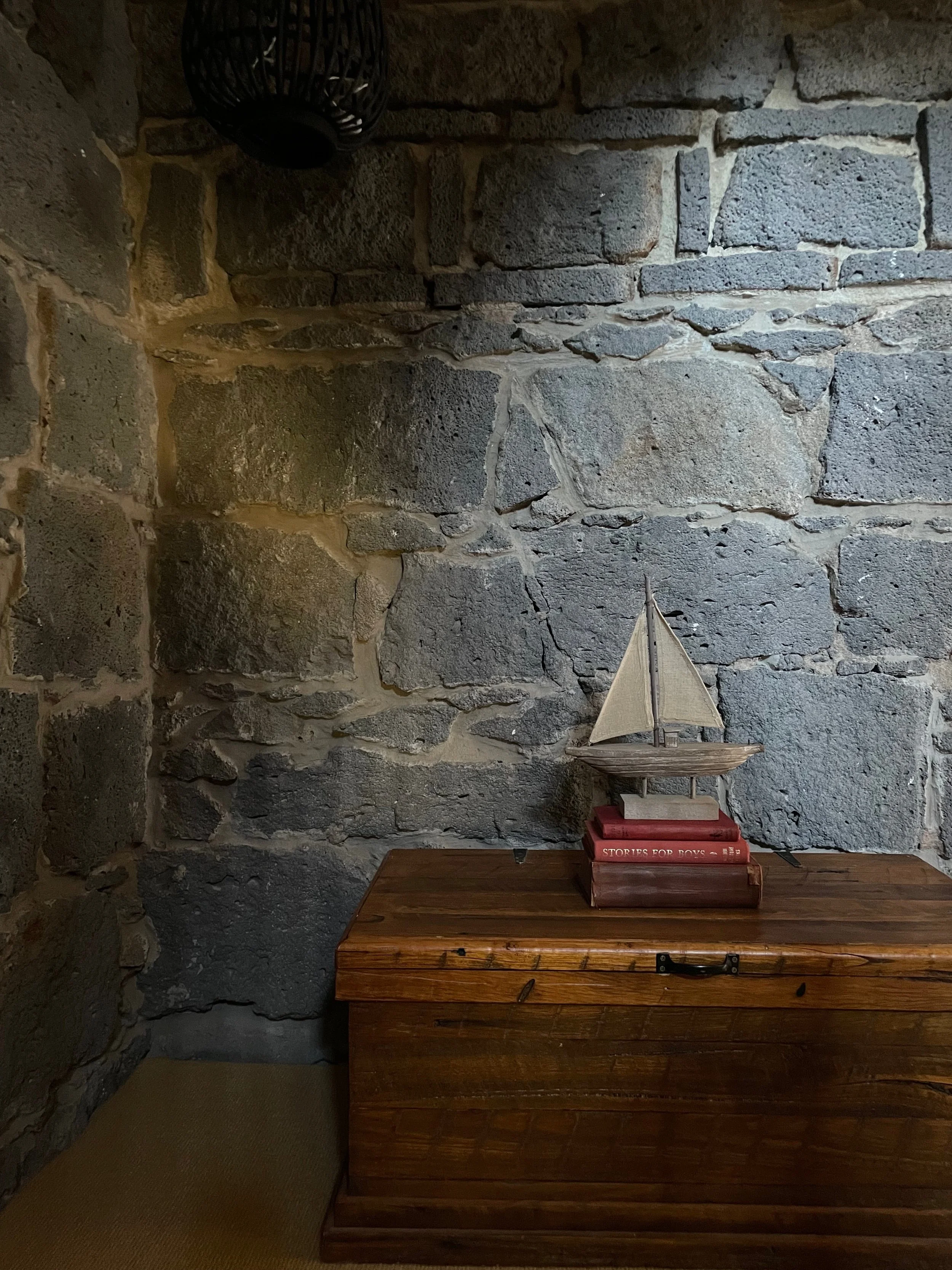 A toy ship sits on vintage books atop a wooden chest. It is surrounded by a rough bluestone wall.