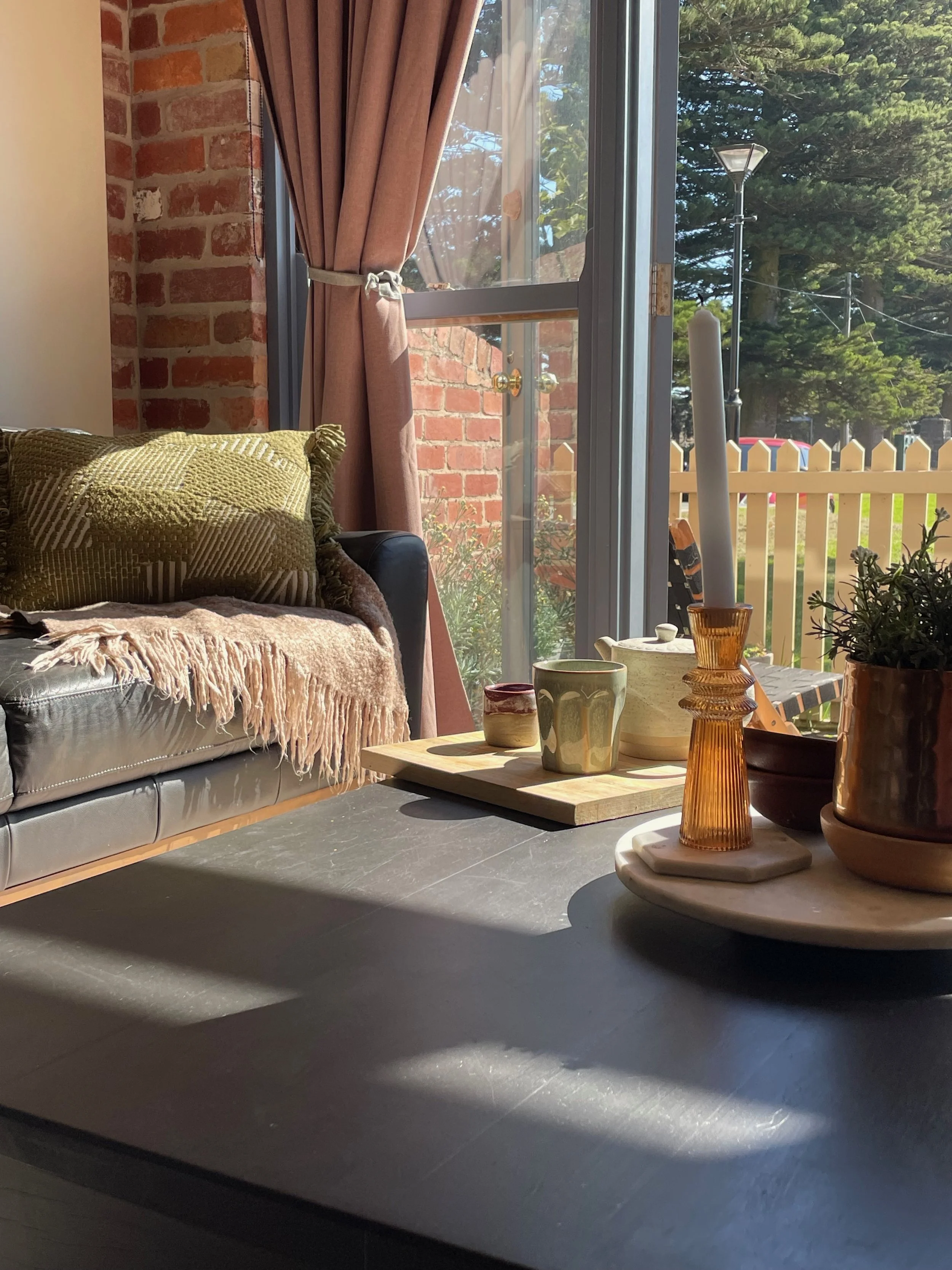 Sunlit living room with beige sofa, green pillow, and throw blanket. Large window with pink curtains showing outdoor greenery and trees. Wooden coffee table with decorative ceramic and glass items, including vases, cups, and a plant in a pot.