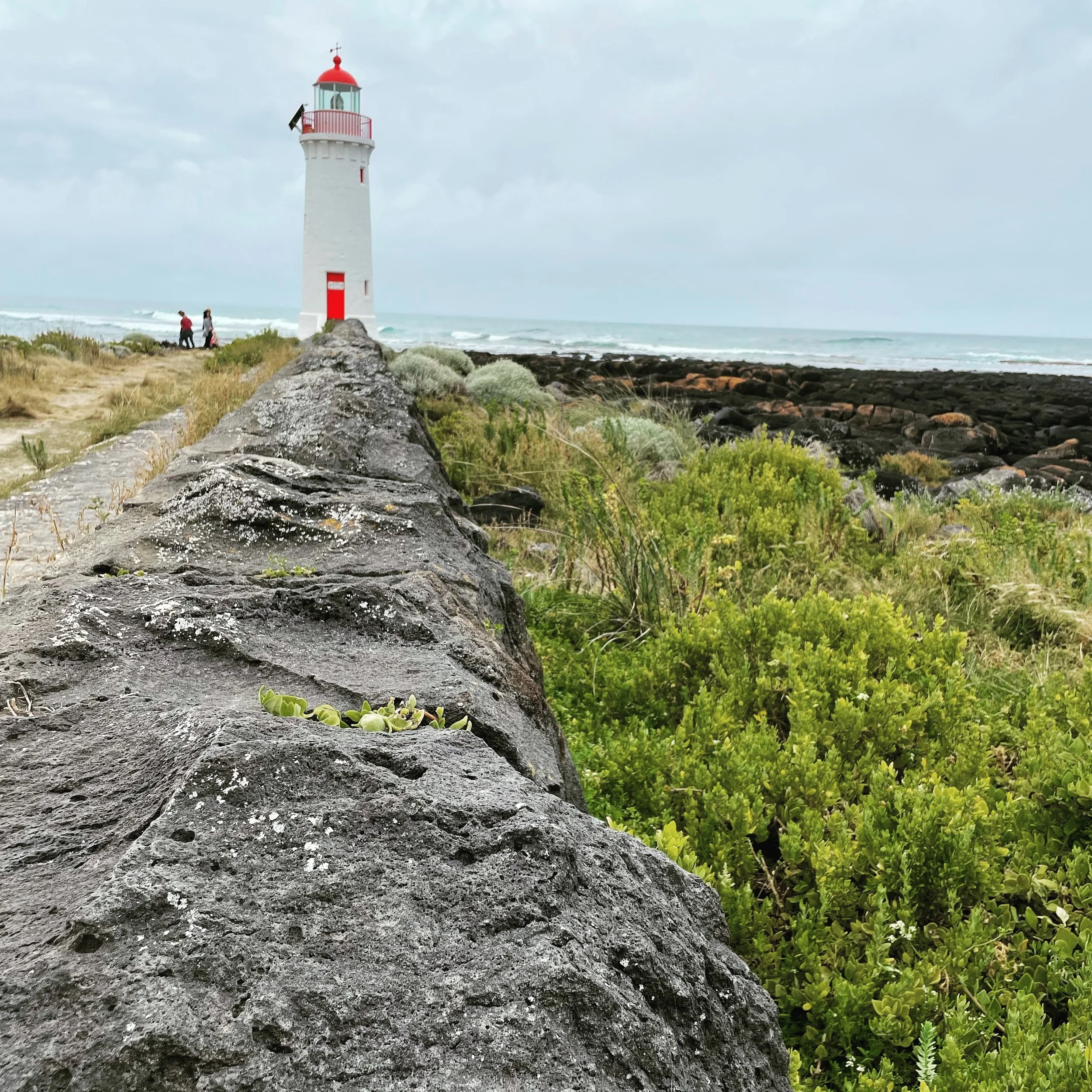 A tall white lighthouse with a red top and railing, located on a rocky shoreline, with two people walking nearby and the ocean waves in the background under overcast sky.