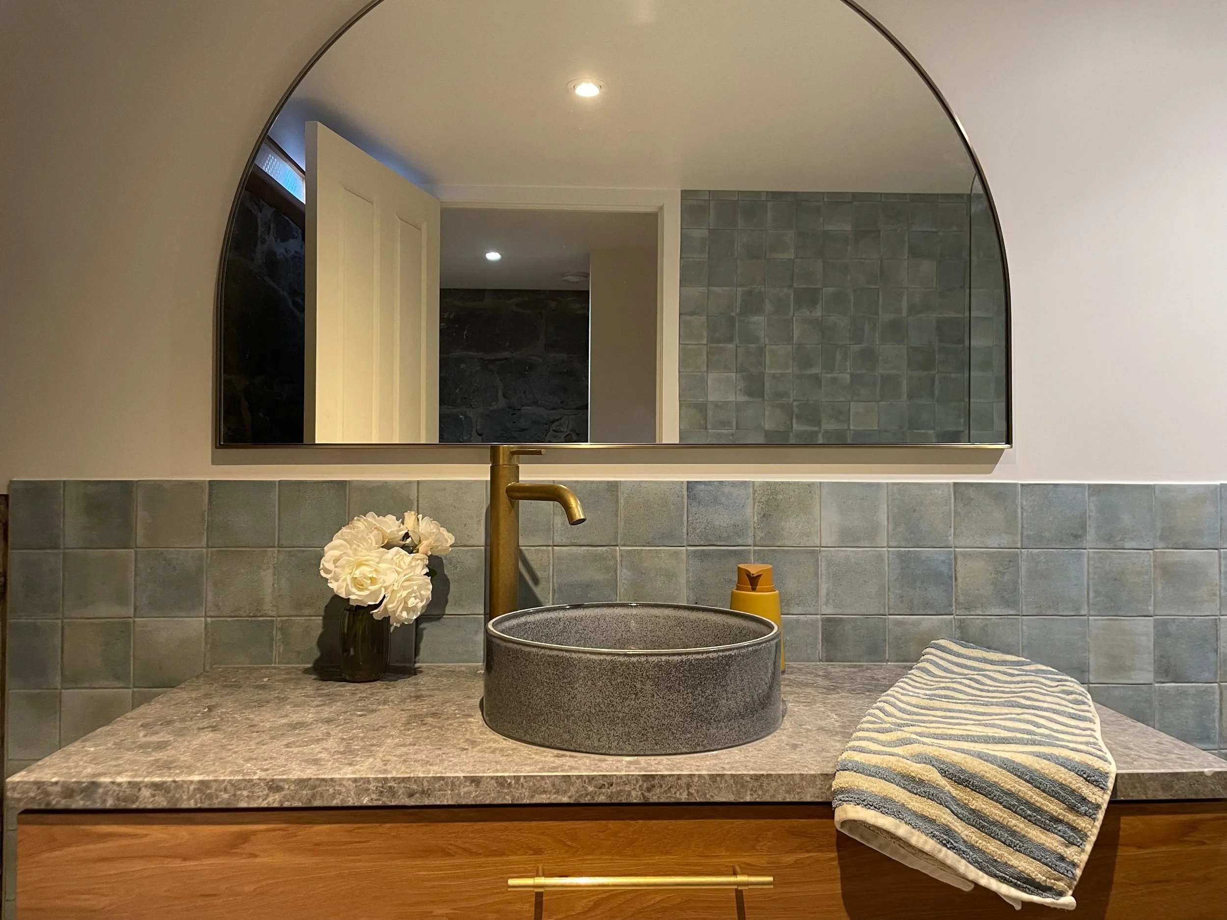 Bathroom with gray tiled backsplash, a large mirror, a white flower arrangement, a textured black basin on the countertop, and a striped towel.