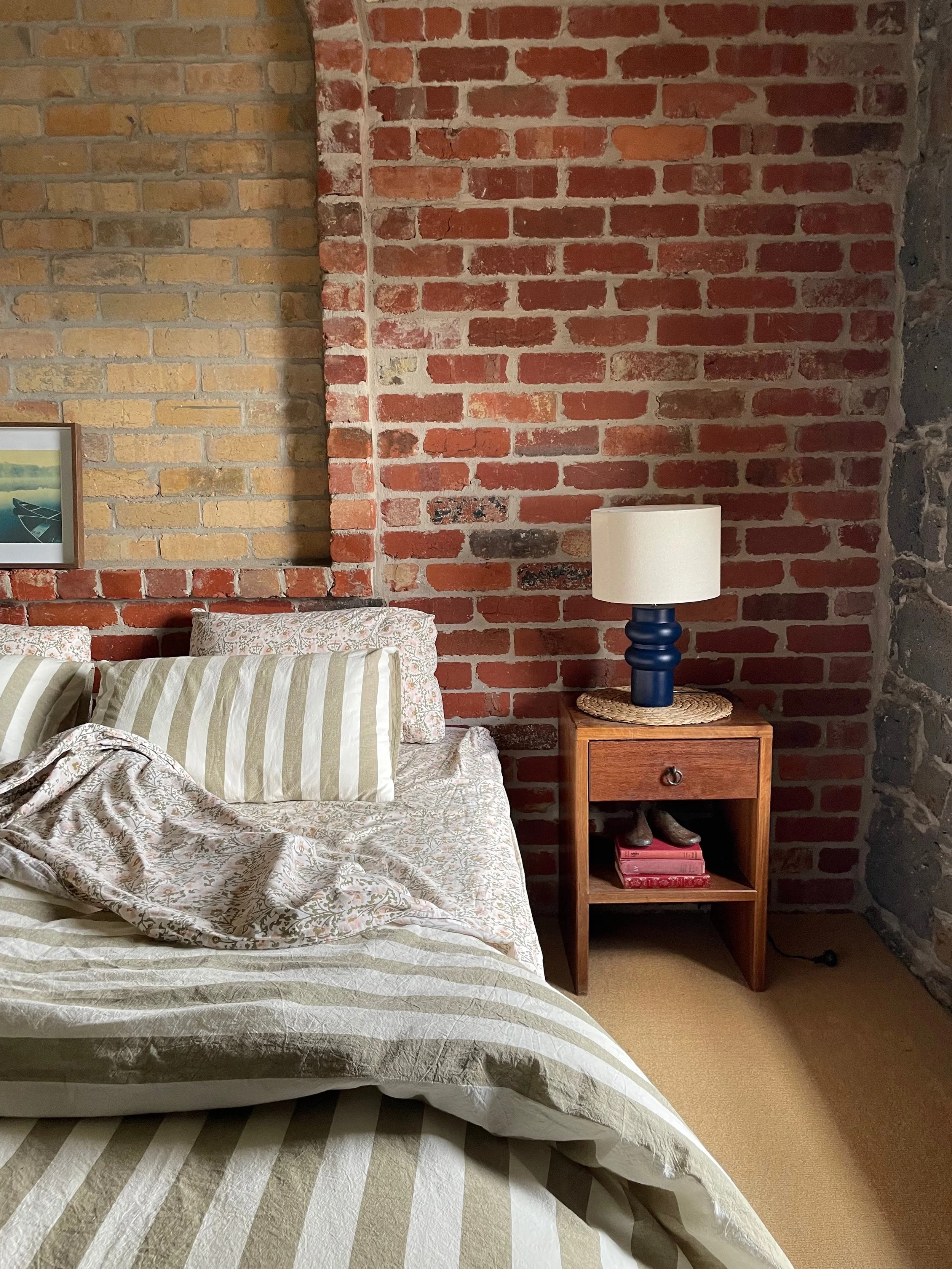 A bedroom with exposed brick walls, a bed with striped bedding, and a wooden nightstand with a lamp and a few books.