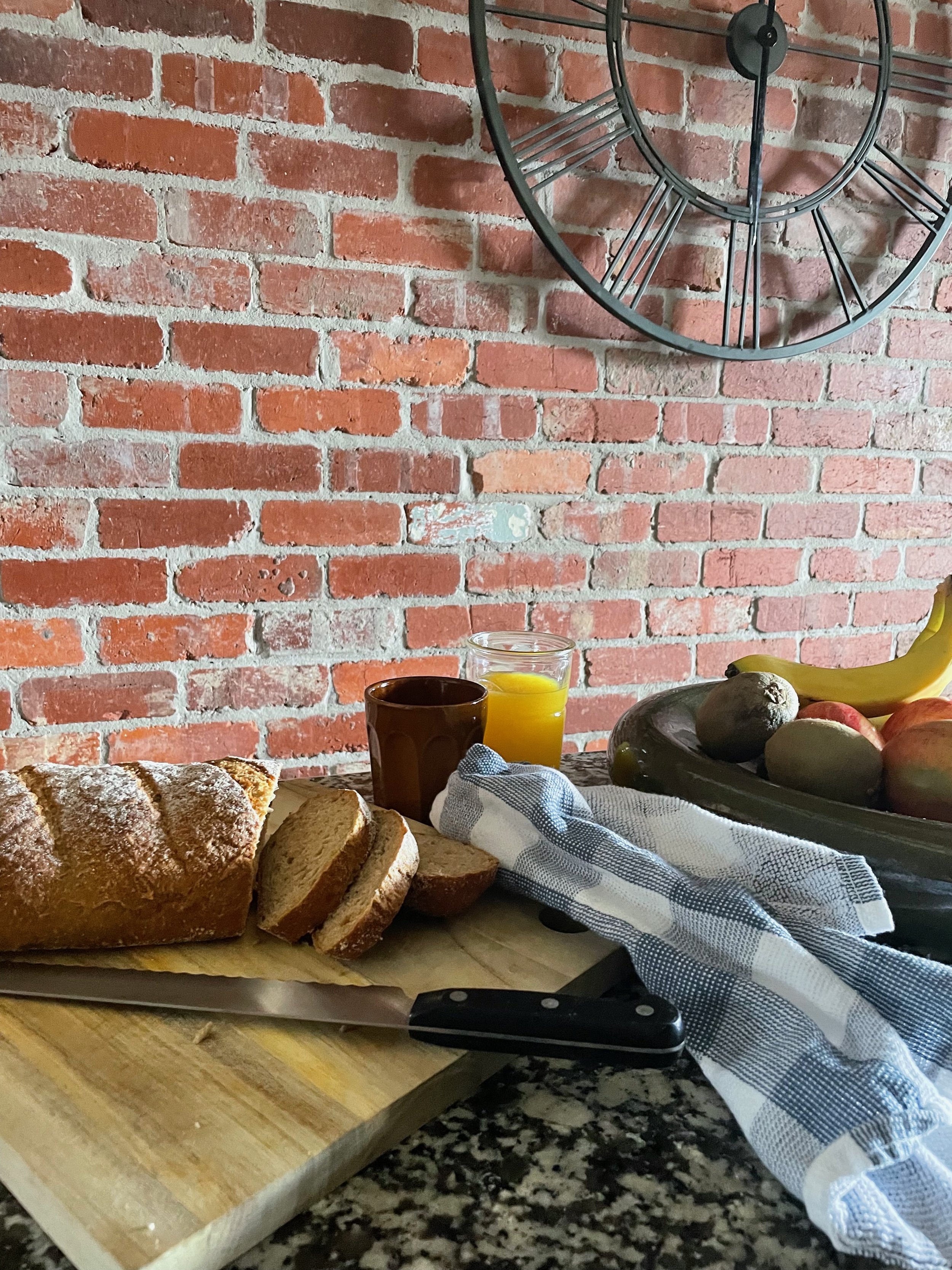 A loaf of sliced bread on a cutting board, with a bread knife on the countertop. In the background, there is a plate of avocados and bananas, a glass of orange juice, a cup, and a blue and white checkered cloth. The scene is set against a brick wall 
