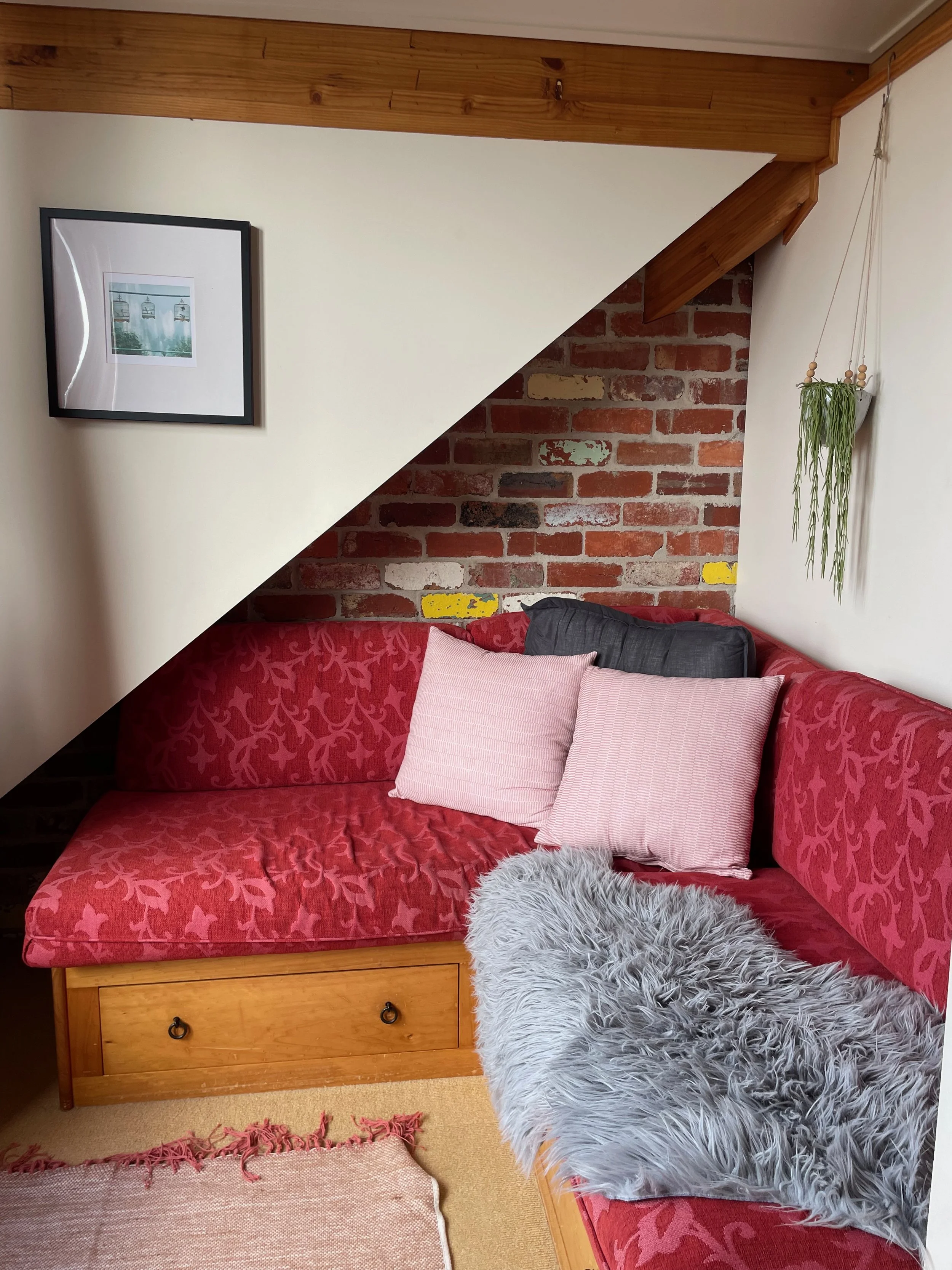 Cozy corner of a room featuring a wooden cabinet beneath a red patterned sofa, with pink and gray pillows, a fluffy gray throw, a brick wall, framed picture, hanging plants, and a section of a carpeted floor.