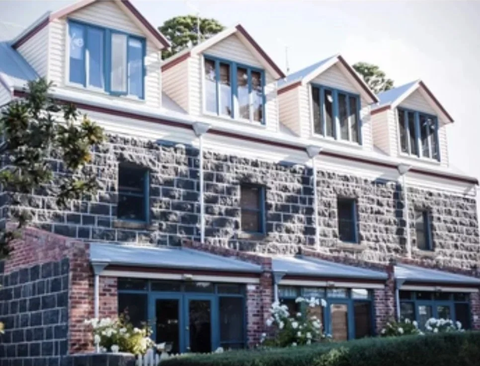 A multi-story house with a stone and brick facade, multiple large windows, and small gabled dormer windows on the roof. There are some plants and flowers in front of the house.