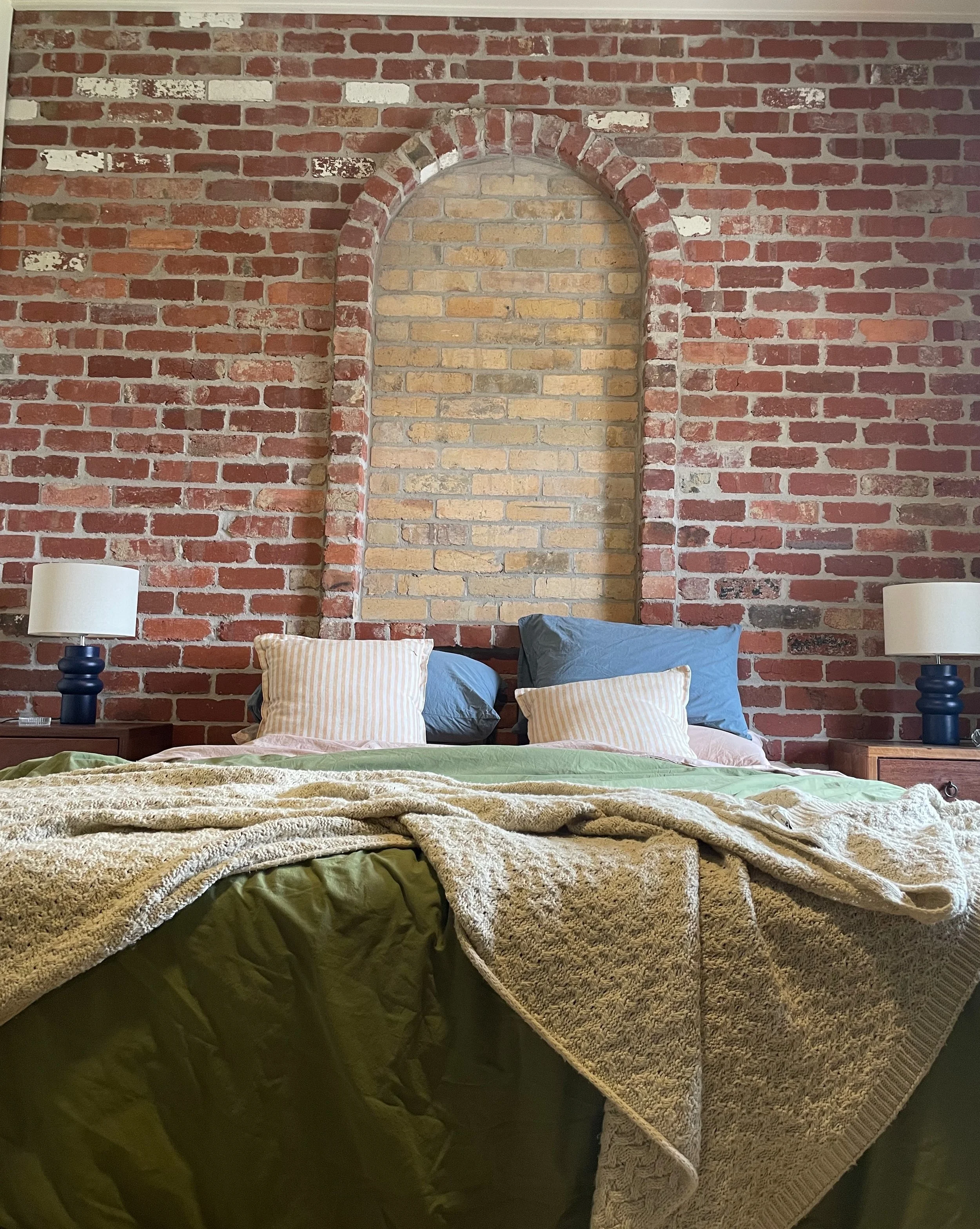 A bedroom with a brick wall headboard, featuring an arched inset in the brick wall, flanked by two lamps on dark blue bedside tables. The bed has various pillows and a blanket, with a green bedspread.
