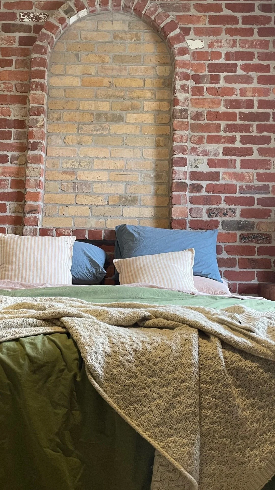 A messy bed in greens, blues and pinks sits in front of a red brink wall, with feature arch of yellow brick.