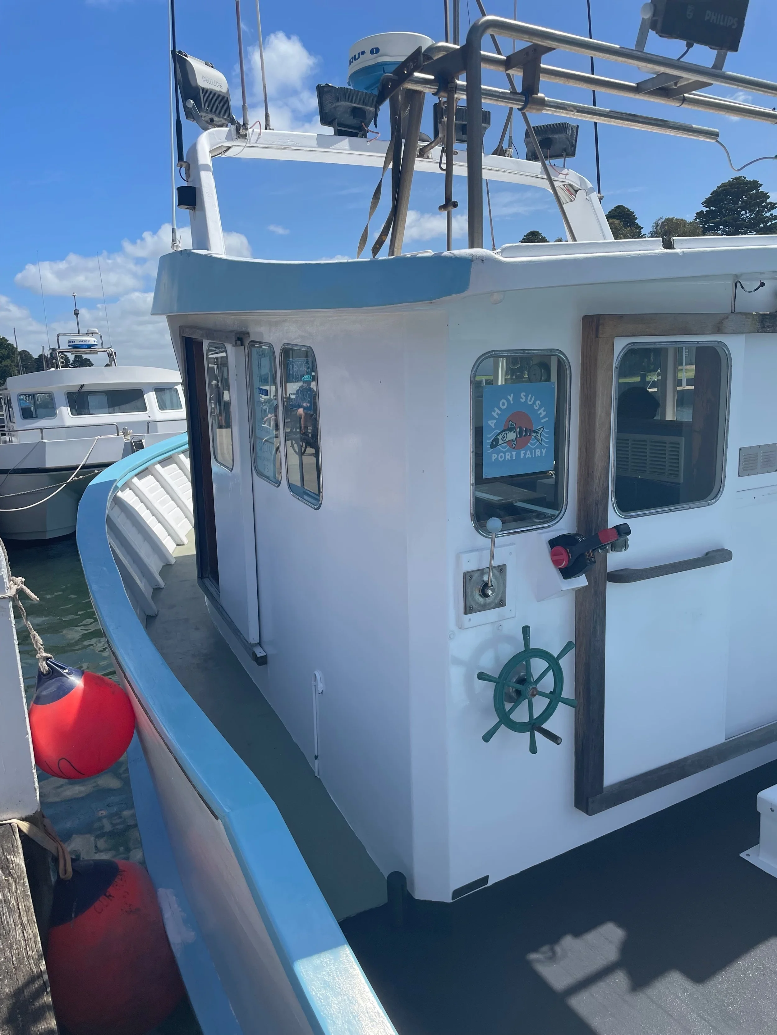 White boat docked at a marina with a red buoy hanging from its side, blue sky with some clouds above, and trees in the background. The boat has a small cabin with windows and equipment on top, including a radar. The boat features a blue and white log