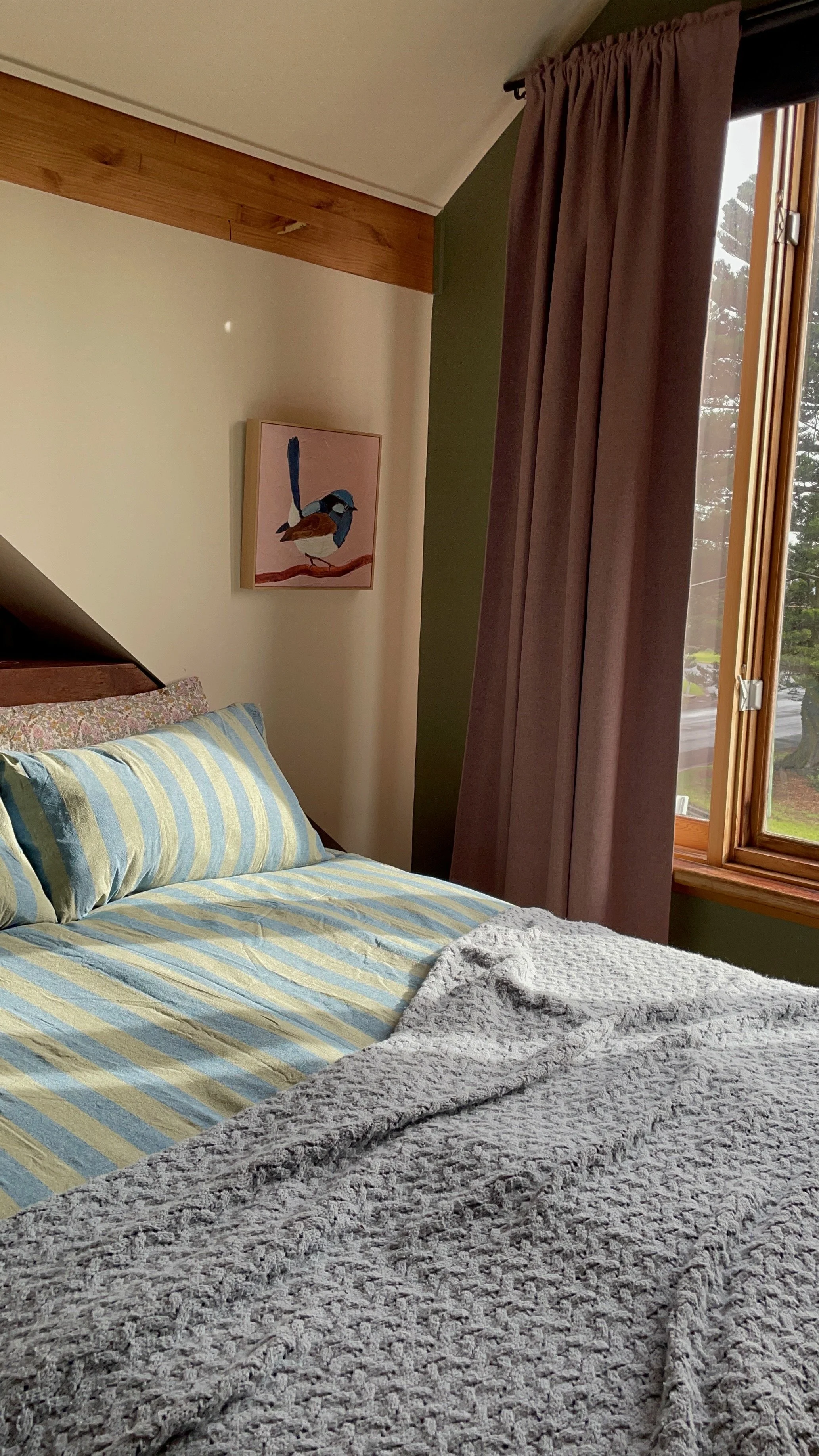 A bedroom with a bed covered in a gray textured blanket, painted yellow and green walls, a window with pink curtains, a framed picture of a bird on the wall, and part of a wooden ceiling.