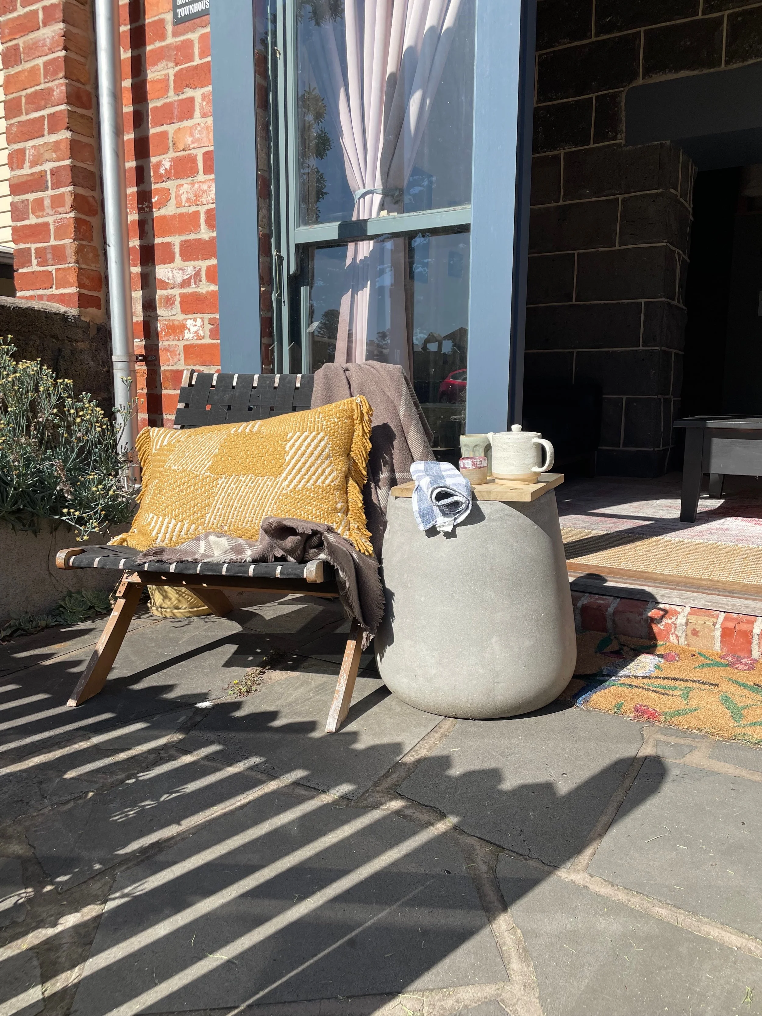 An outdoor patio with a black chair, yellow pillow, gray throw blanket, and a small wooden table holding a ceramic pitcher, mug, and small bowl. There's a large concrete planter, a patterned rug, a glass window with white curtains, and brick and dark wall structures with shadows cast on the ground.