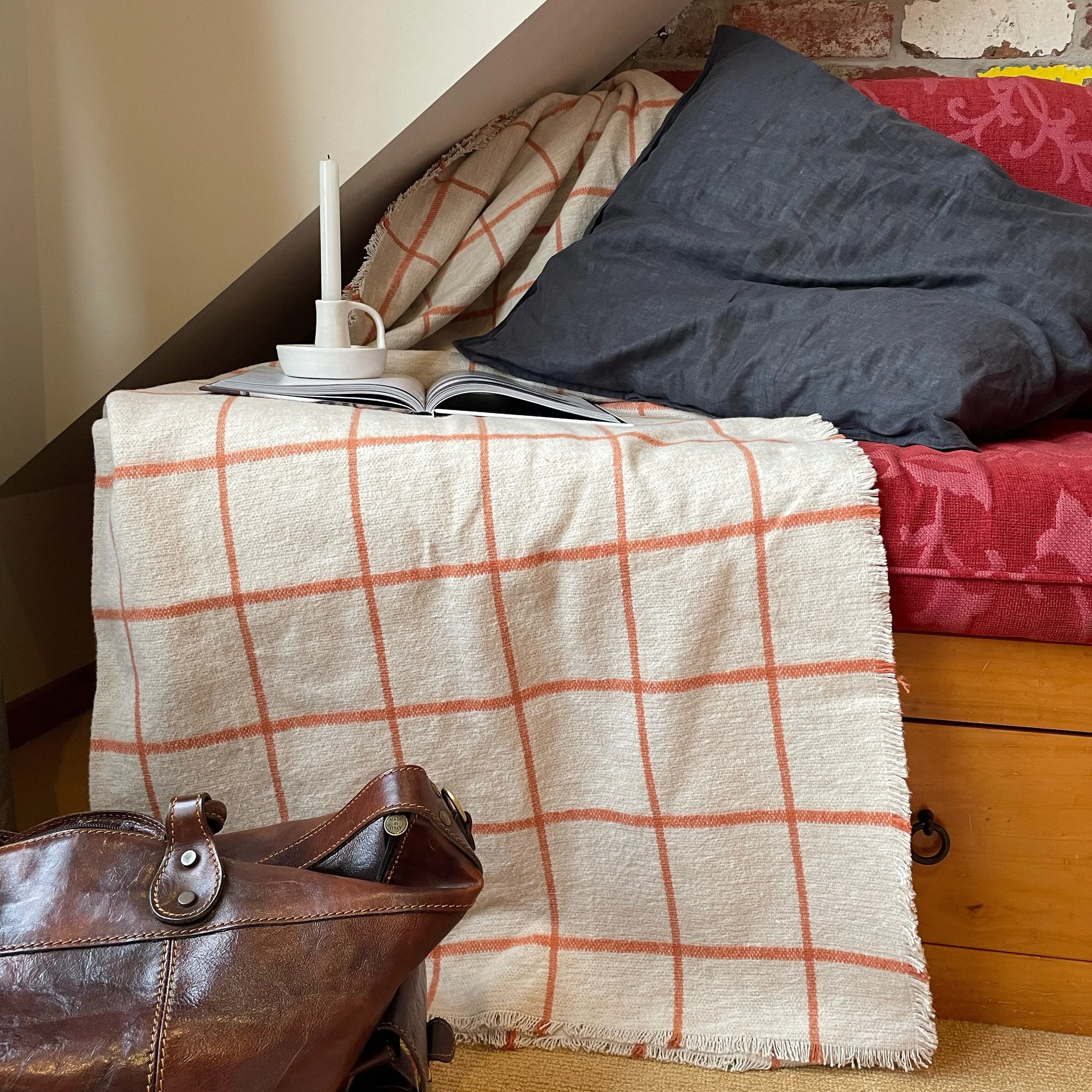 A bed with a plaid blanket, a gray pillow, an open book, and a white candle holder shaped like a candle, against a brick and painted wall.