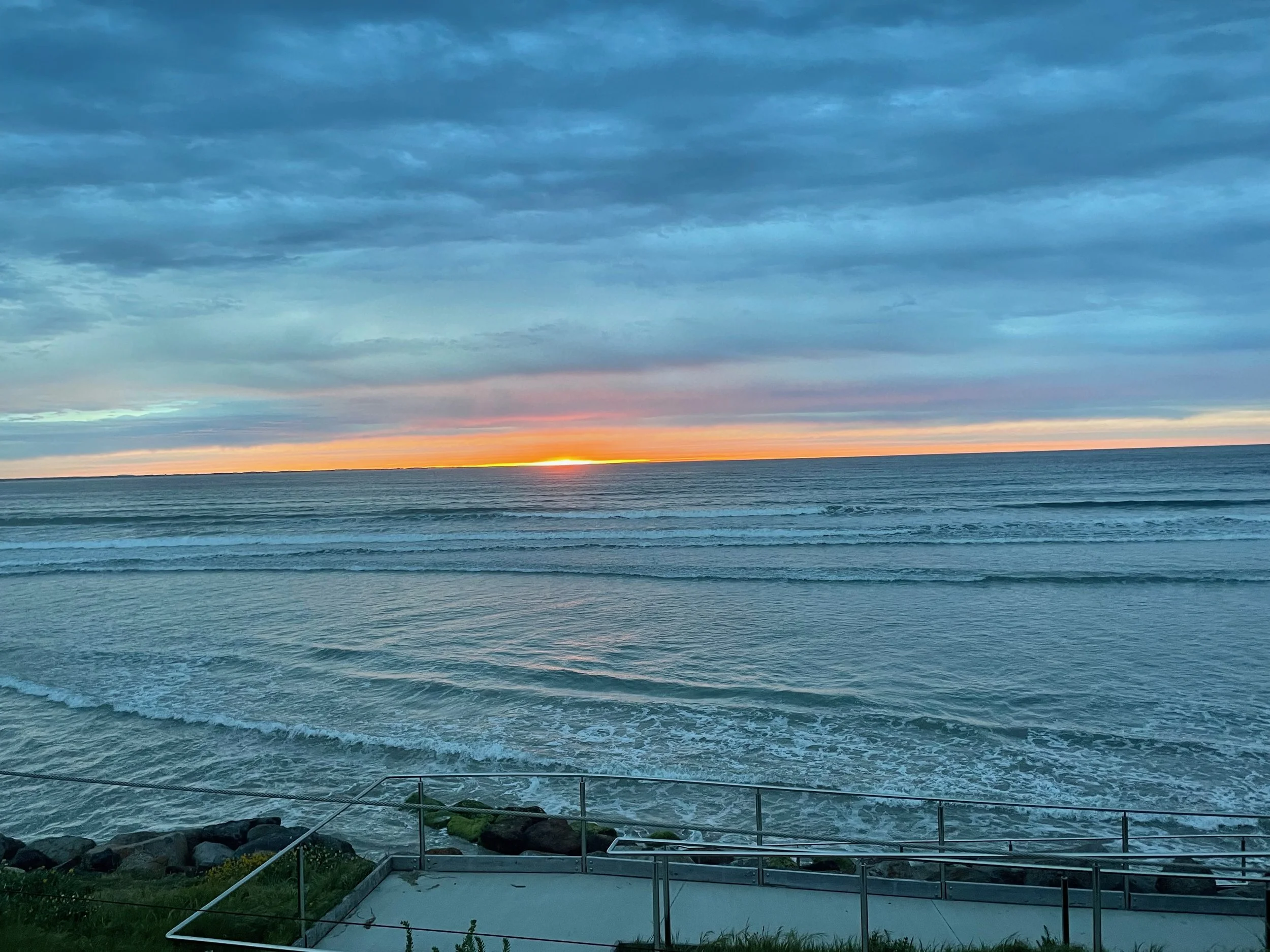 View of the ocean at sunset with a partly cloudy sky, waves crashing, and a shoreline with rocks and a pathway in the foreground.