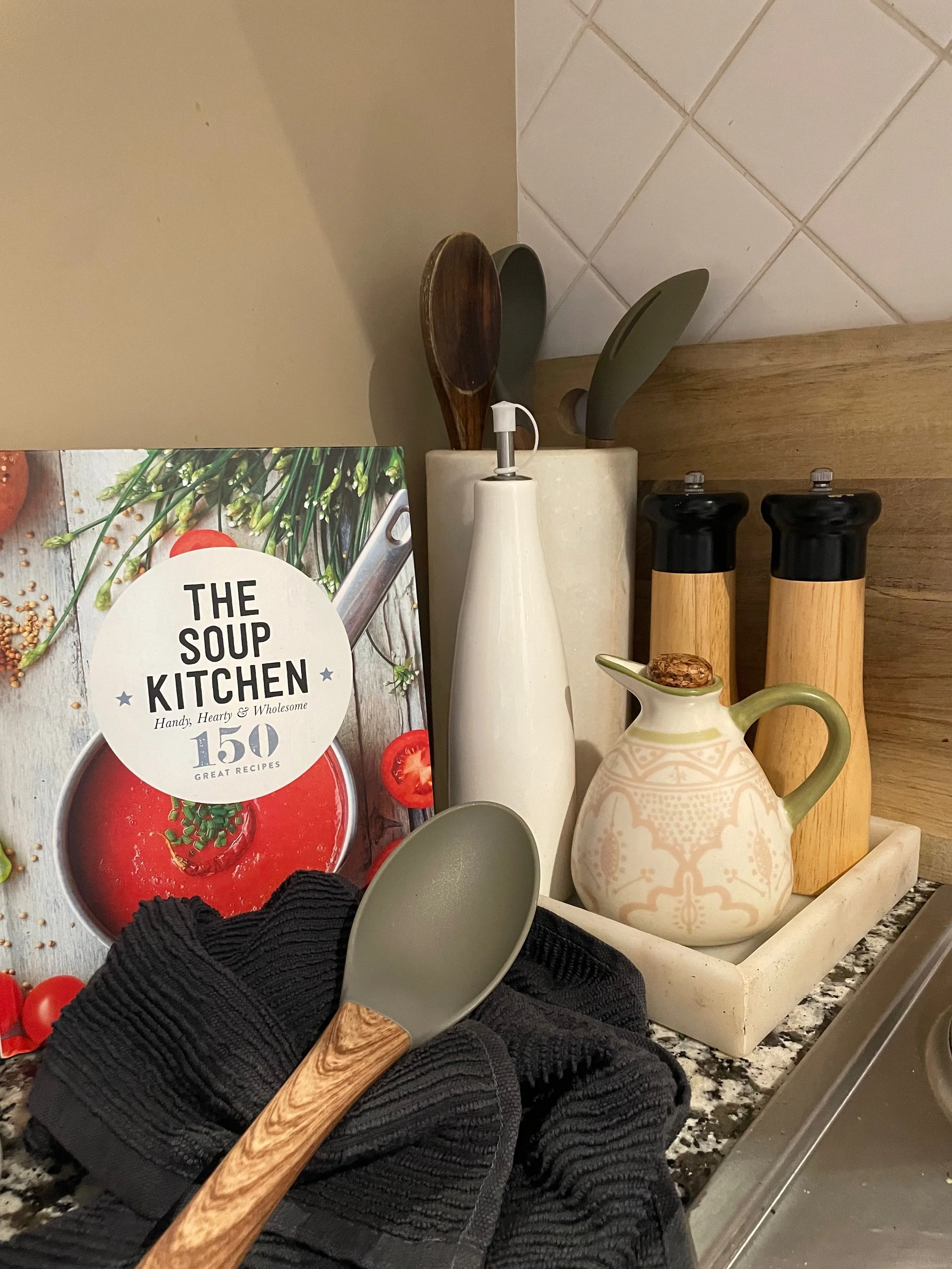 A kitchen countertop with a black cloth, a spoon with a wooden handle, and a person wearing black gloves holding the spoon. In the background are a cookbook titled 'The Soup Kitchen,' salt and pepper shakers, and a decorative pitcher.