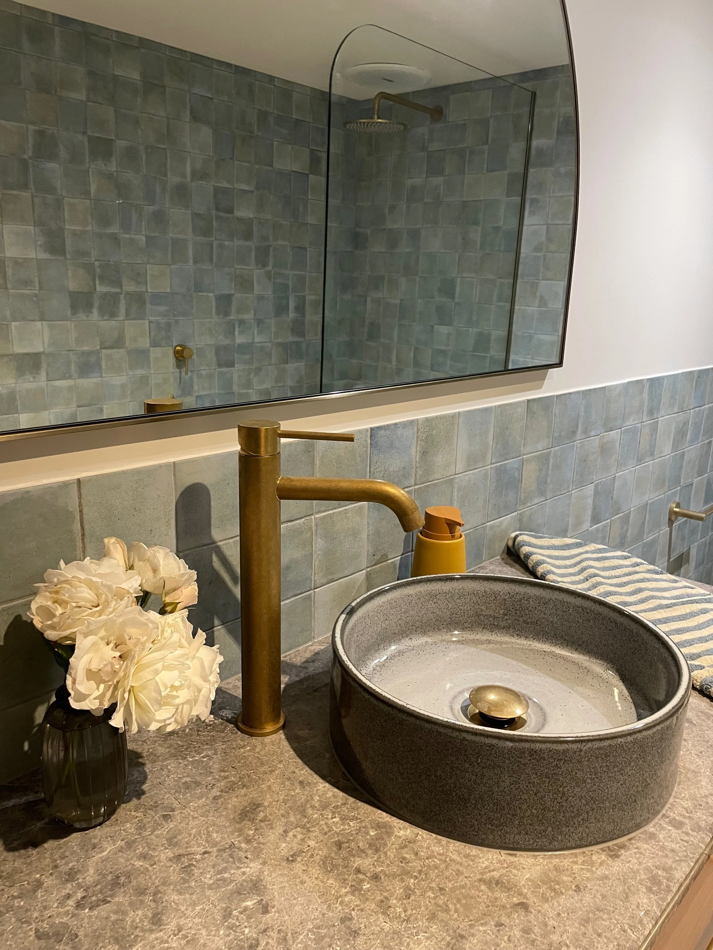 A bathroom countertop with a black vase holding white flowers and a tan soap dispenser. A round, dark gray sink with a gold drain is in the foreground. There is a gold faucet and a striped towel on a ledge. A large rectangular mirror above reflects a