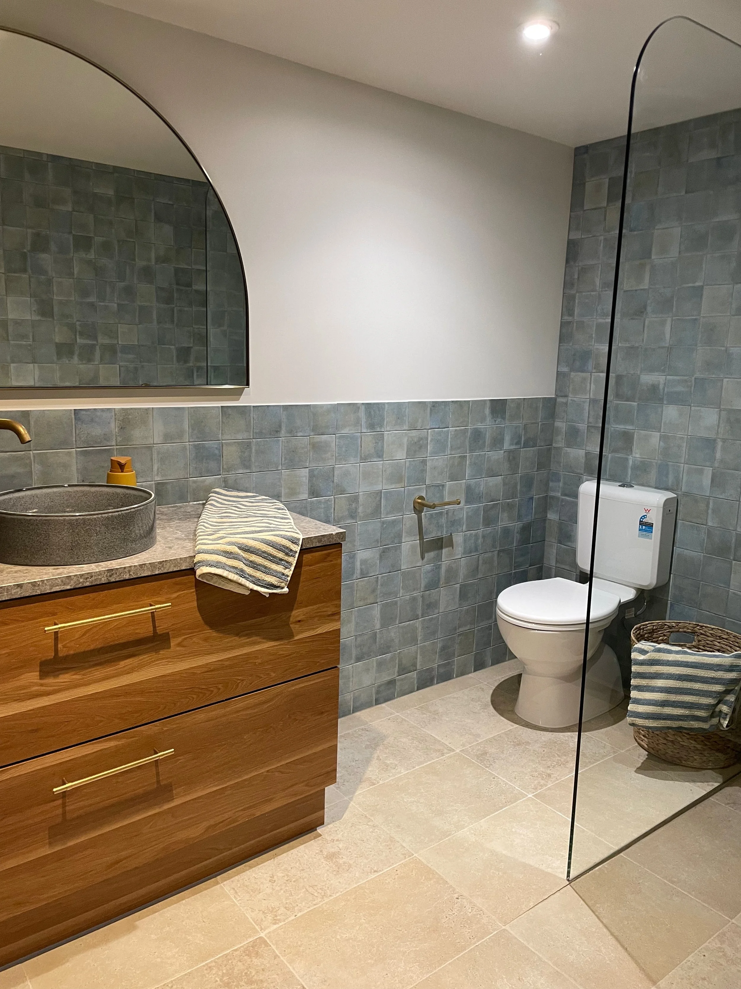 A modern bathroom with a wooden vanity, round dark basin, and striped towel. There is a mirror, a toilet, and a wicker basket, with blue-gray tiled walls and beige floor tiles.
