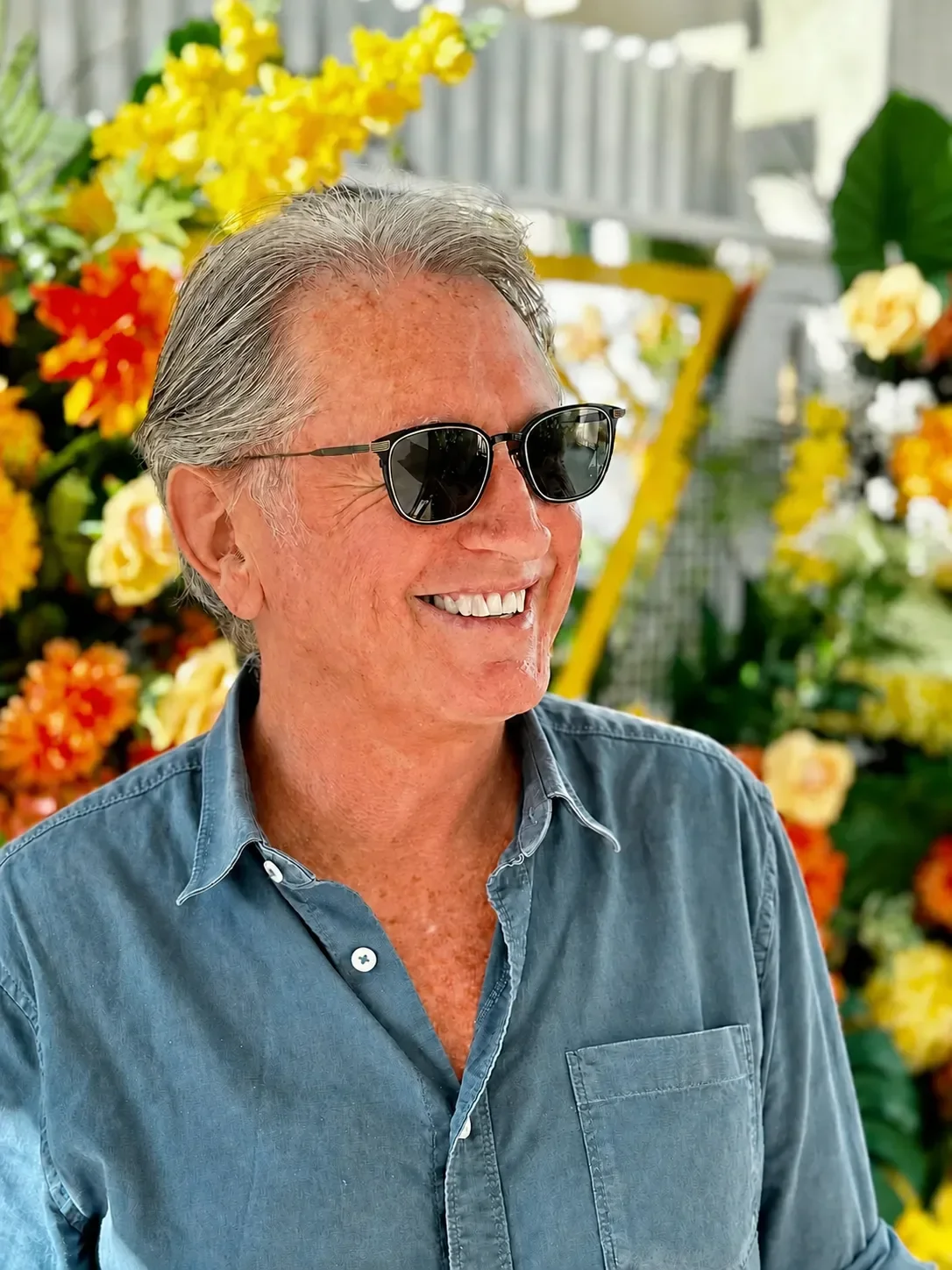 Portrait of Brendan Callinan, author, musician and business consultant, seated in front of orange and yellow flowers wearing black sunglasses.