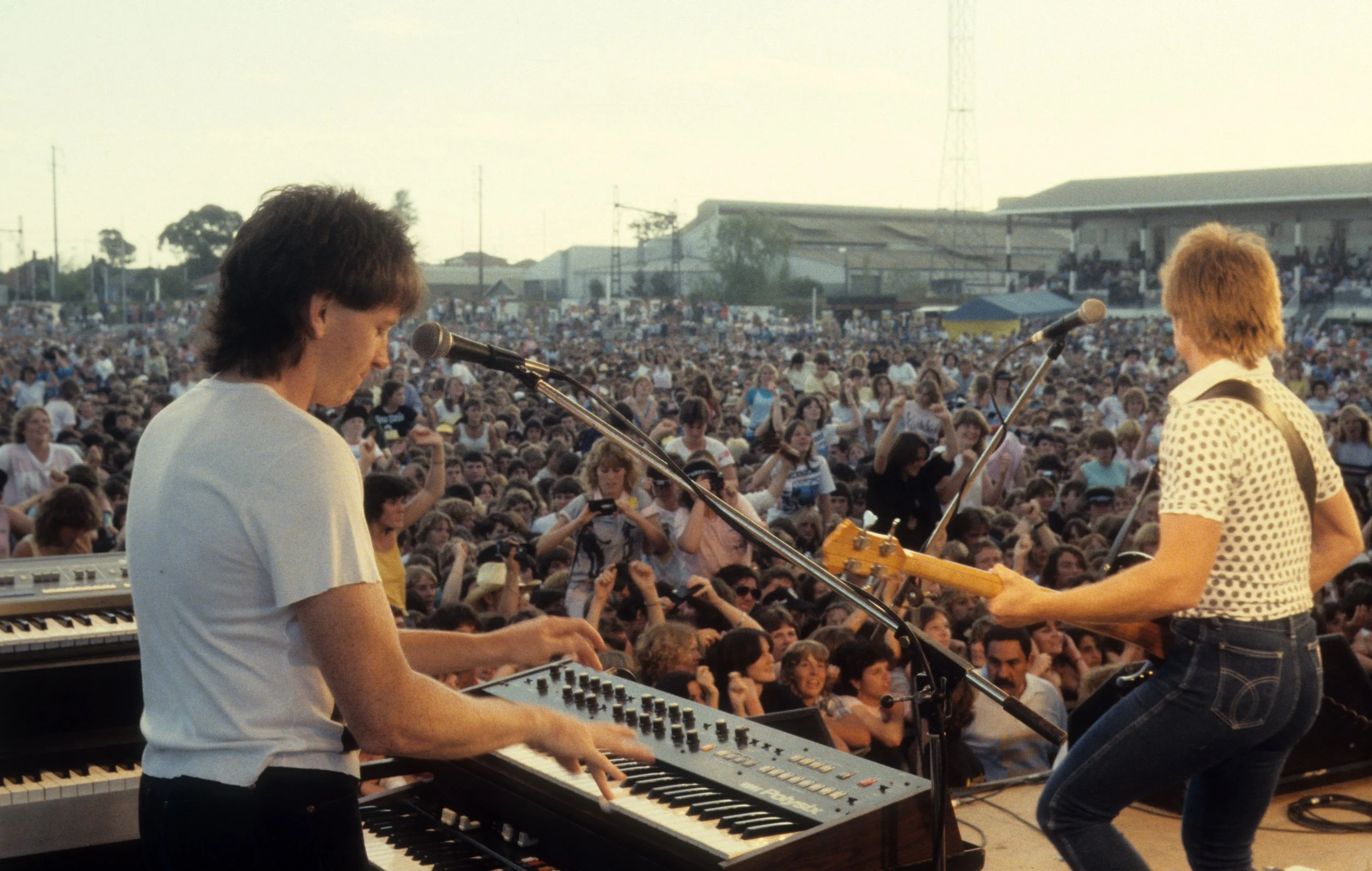 Two musicians performing on an outdoor stage in front of a large crowd. One is playing keyboard and the other guitar in the iconic Aussie band, The Radiators.