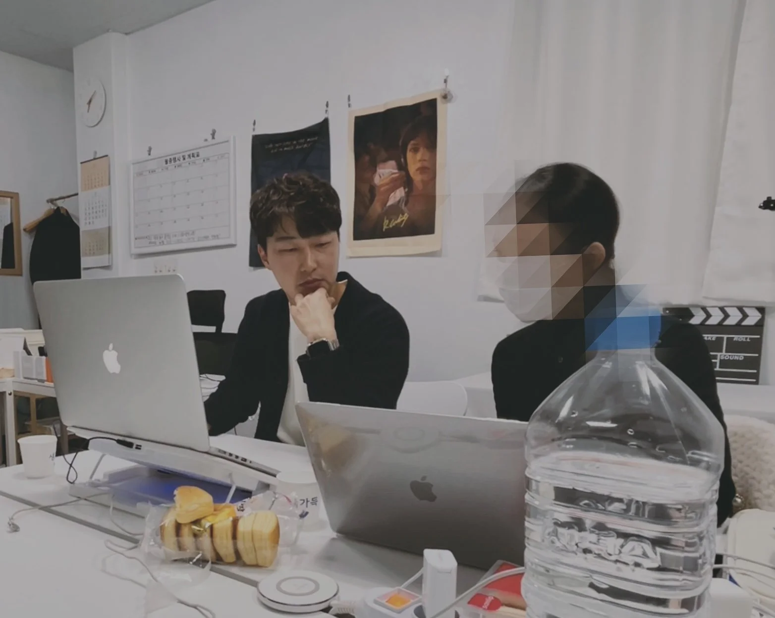 회의실에서 두 명의 남성이 노트북과 대화 중이며, 탁자에 먹거리와 물병이 놓여 있음.