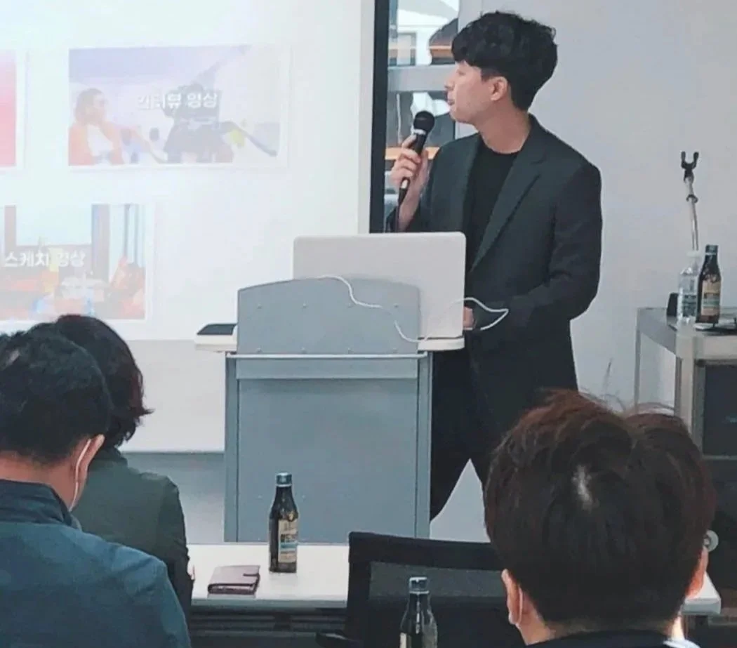 남자가 마이크를 들고 프레젠테이션을 하고 있으며, 뒤에 프로젝터 화면에 여러 이미지와 한국어 텍스트가 보여지고 있다. 참석자들이 강의를 듣고 있다.